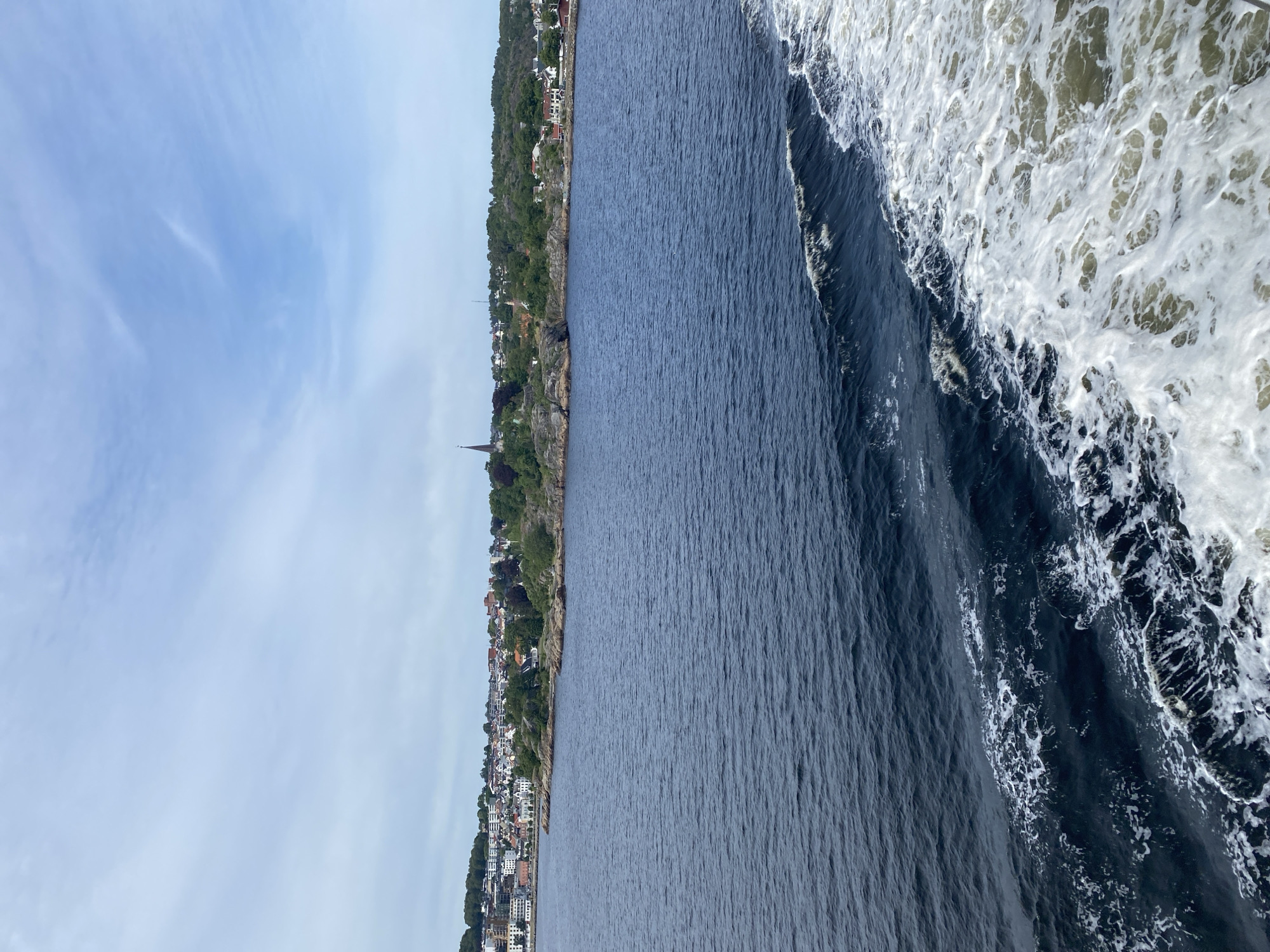 This is a vertical, medium-shot photograph taken from a boat moving through a body of water, likely a fjord or coastal area, near Larvik, Norway. The scene depicts a scenic view of a waterfront town nestled along a lush, green, and hilly coastline. The town is characterized by clusters of white and red-roofed buildings, with a prominent church steeple visible among them. The water in the foreground is dark blue, with white foam and waves indicating the boat's movement. The sky is a pale blue with wispy clouds. The time of day appears to be daytime, and the weather seems pleasant. The overall mood is serene and picturesque, highlighting the natural beauty of the Norwegian coast and its charming settlements. There is no visible text in the image.