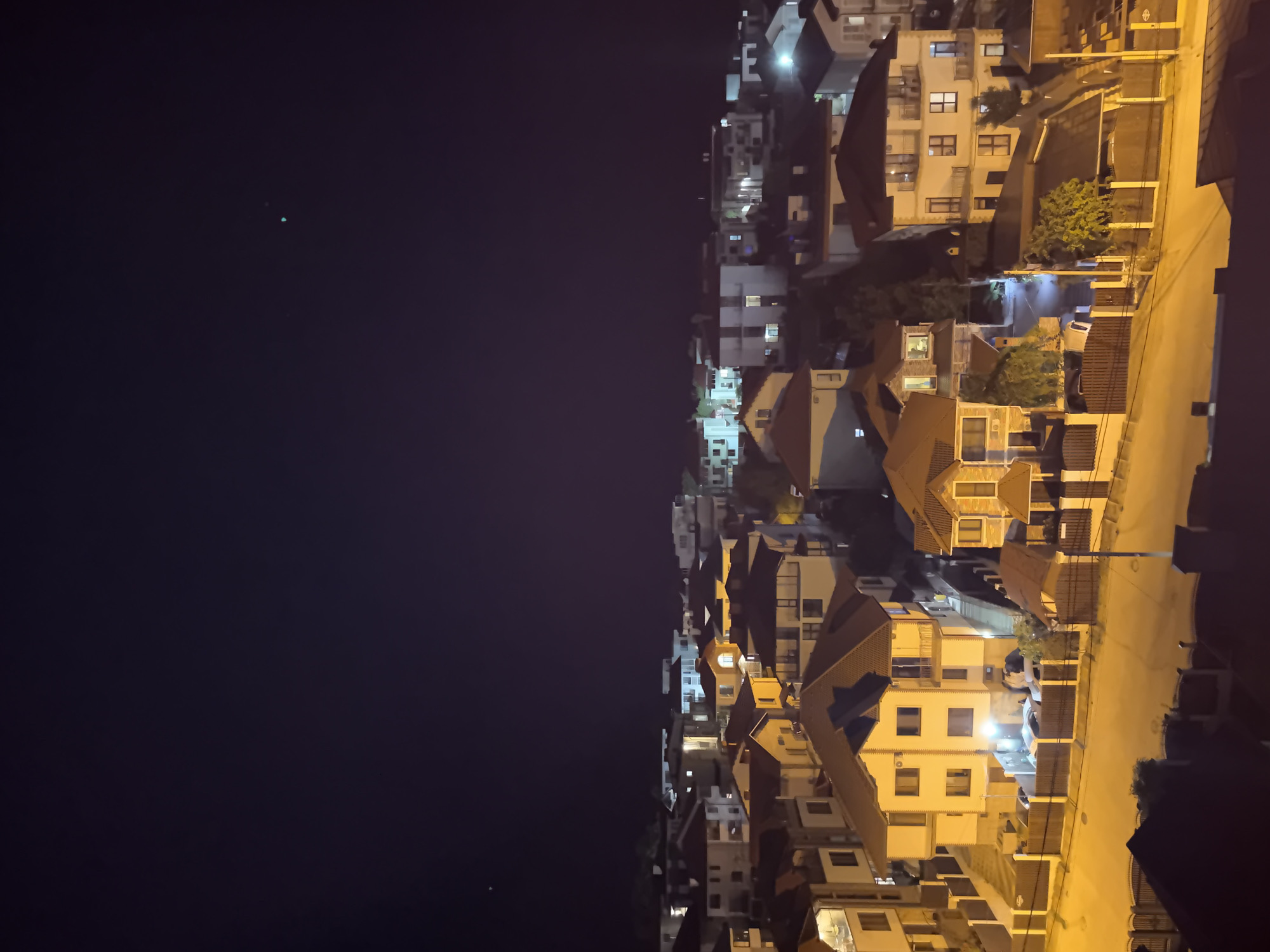 This is a nighttime aerial view of a residential area, likely on a hillside, with multiple houses and buildings visible. The sky is dark, with two small, bright points of light visible, possibly stars or distant lights. The houses are illuminated from within by yellow and white lights, casting a warm glow. Many of the houses have red-tiled roofs and feature balconies and multiple stories. Streets and pathways are visible between the houses, illuminated by streetlights. Fences, some made of wood and others of metal, surround many of the properties. In the foreground, several rooftops are visible, with one appearing to be a dark-tiled roof with a lighter colored edge. The overall scene depicts a quiet, urban residential setting at night.