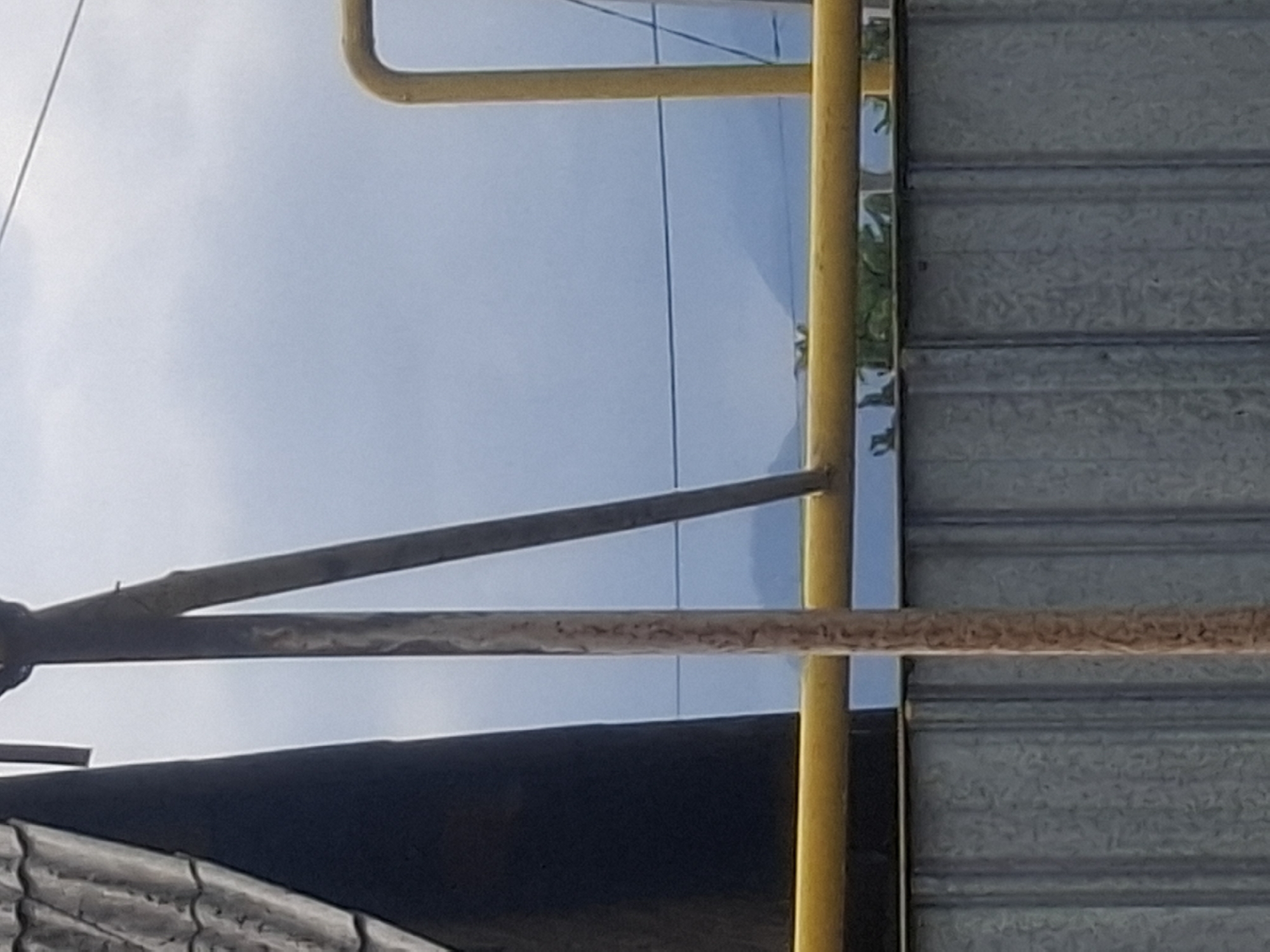 The image captures a portion of an outdoor scene, likely in Almaty, Kazakhstan, focusing on structural elements and the sky. In the foreground, a corrugated metal fence runs horizontally, appearing to be a boundary. Above the fence, a bright yellow pipe or conduit is visible, also running horizontally. To the left, a dark, conical structure with a tiled roof suggests a building or a silo. A vertical, weathered wooden pole stands prominently in the center, with a metallic pole behind it. The background consists of a hazy sky with faint outlines of distant mountains, indicating a potentially overcast or smoggy day. There are no people or discernible actions taking place in the frame. The overall impression is one of utilitarian infrastructure against a natural, albeit obscured, landscape.