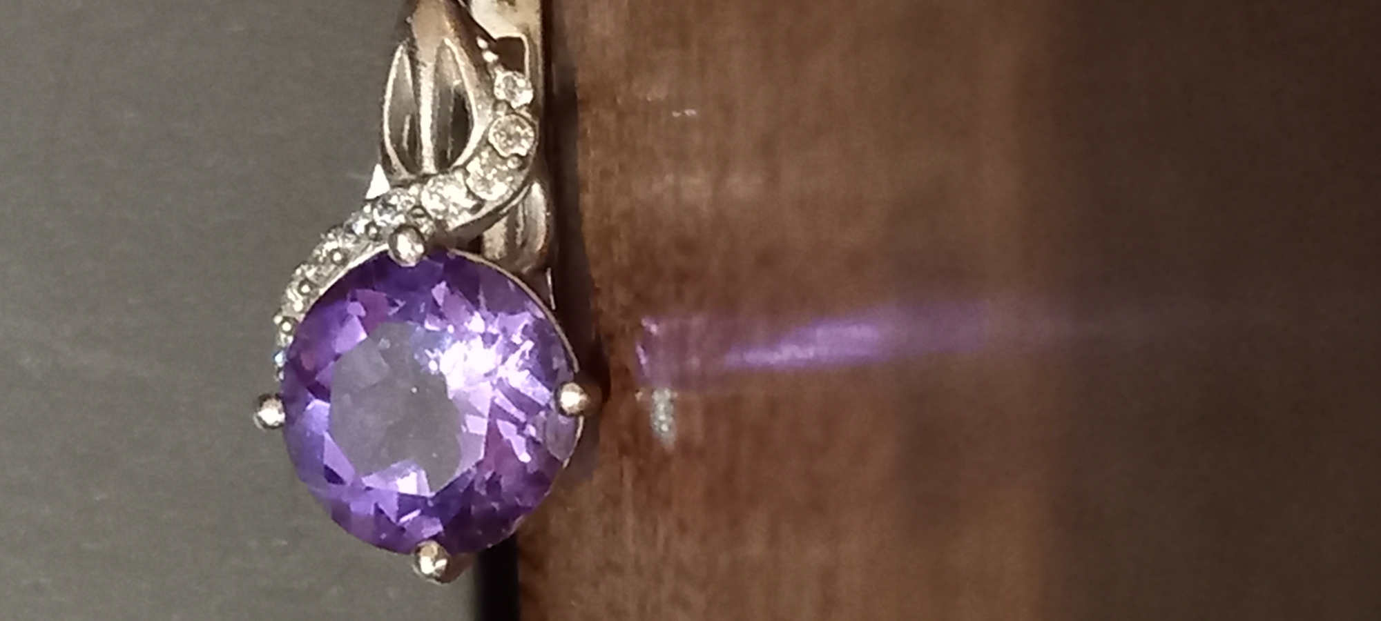A close-up, still life photograph displays a single piece of jewelry, likely an earring or pendant, against a split background. The main subject is a large, round, faceted purple gemstone, most likely an amethyst, held by four prongs in a silver-colored metal setting. Above the main stone, the setting features an intricate, possibly twisted or interwoven design, adorned with numerous small, clear, sparkling stones, suggestive of diamonds or cubic zirconia.
The jewelry item is positioned against two distinct backgrounds. To the left, there is a plain, dark gray or black surface. To the right, a warm, reddish-brown background with a subtle texture is visible, possibly wood. A noticeable purple light reflection or glare emanates from the main gemstone onto this brown surface, highlighting its vibrant color and the interplay of light.
The focus is sharp on the jewelry, emphasizing the cut and brilliance of the stones, while the backgrounds are slightly blurred. No human subjects or activities are depicted. There is no visible text in the image. The lighting appears direct, enhancing the sparkle of the smaller stones and the facets of the larger purple gem. No cues for time of day, weather, or specific location are present within the image itself.