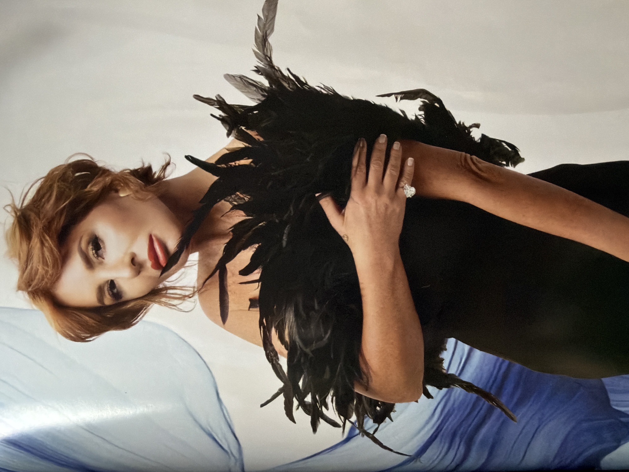 A woman with red lipstick and light brown hair is lying on her side, wearing a black dress with black feathers around her shoulders. She has a large diamond ring on her left hand, which is raised near her shoulder. A blue drape is visible in the lower left of the frame, suggesting a soft, textured surface. The background is a soft white. There is no indication of Moscow, Russia, in the image.