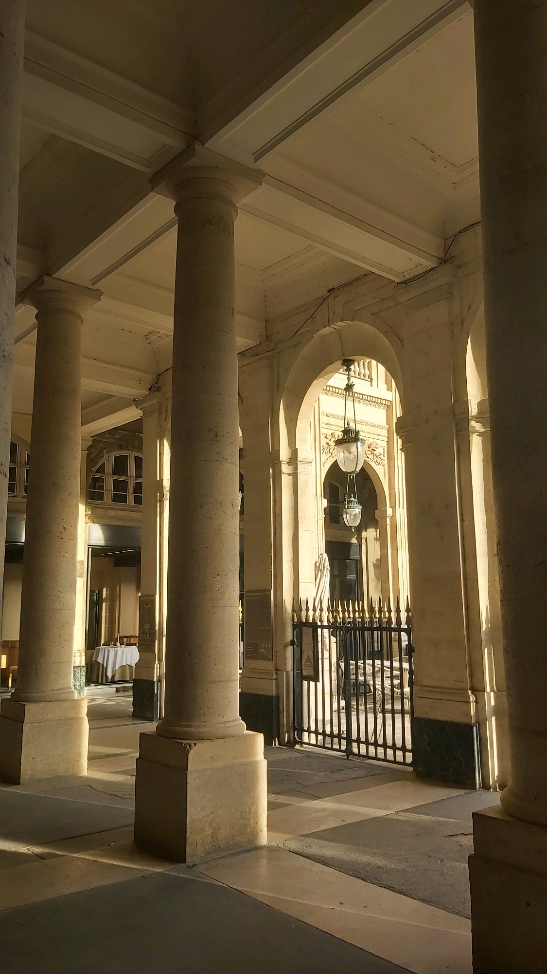 The image shows a sun-drenched colonnade with large stone columns and arched walkways, characteristic of Parisian architecture. The light casts strong shadows on the patterned floor. In the background, through an archway, a black wrought-iron gate is visible, beyond which are outdoor tables and chairs, suggesting a cafe or restaurant setting. Two ornate hanging lamps, one above the other, are suspended from the ceiling within the arch. To the left, a round table covered with a white tablecloth is set with a chair, hinting at an indoor dining area. The overall scene conveys a sense of classical elegance and a peaceful, sunlit atmosphere in a Parisian location.
