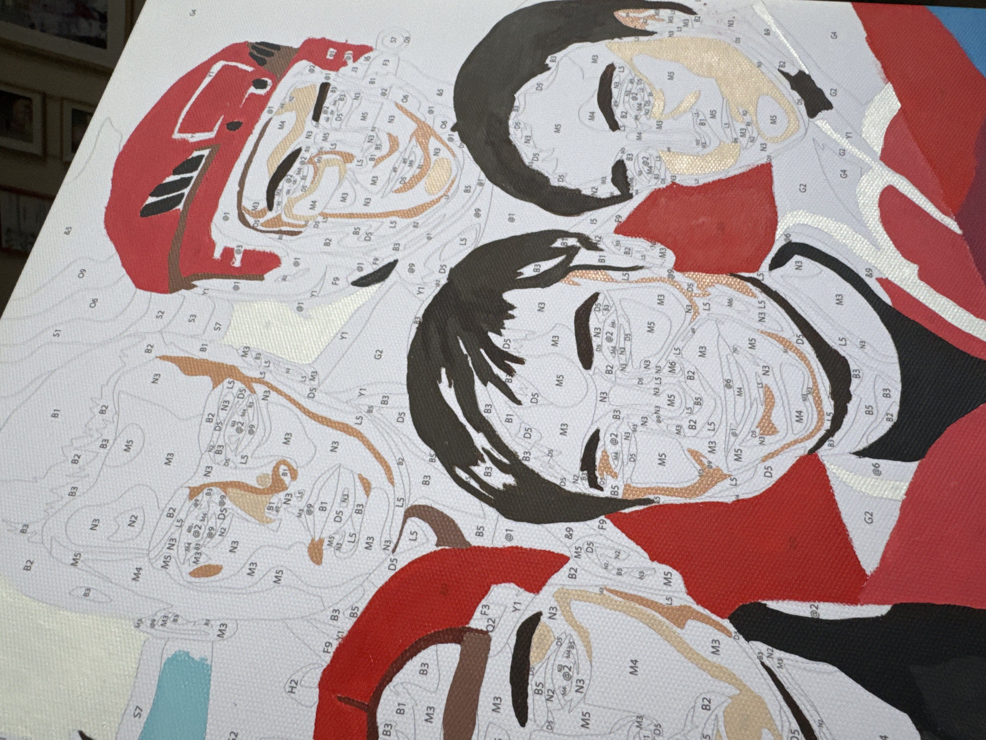 The image shows a paint-by-numbers canvas depicting several people, possibly hockey players, wearing jerseys and helmets. The canvas is filled with numbered sections, indicating the colors to be used. The figures appear to be arranged in a composition of overlapping portraits. The background and some parts of their jerseys are already painted in shades of red, black, white, and gray. The figures' faces and parts of their jerseys remain unpainted, revealing number and letter codes. The scene takes place indoors, suggested by the presence of frames and other objects in the background, which is out of focus. The location in the city of Chaikovsky, Russia is not determinable from the image.