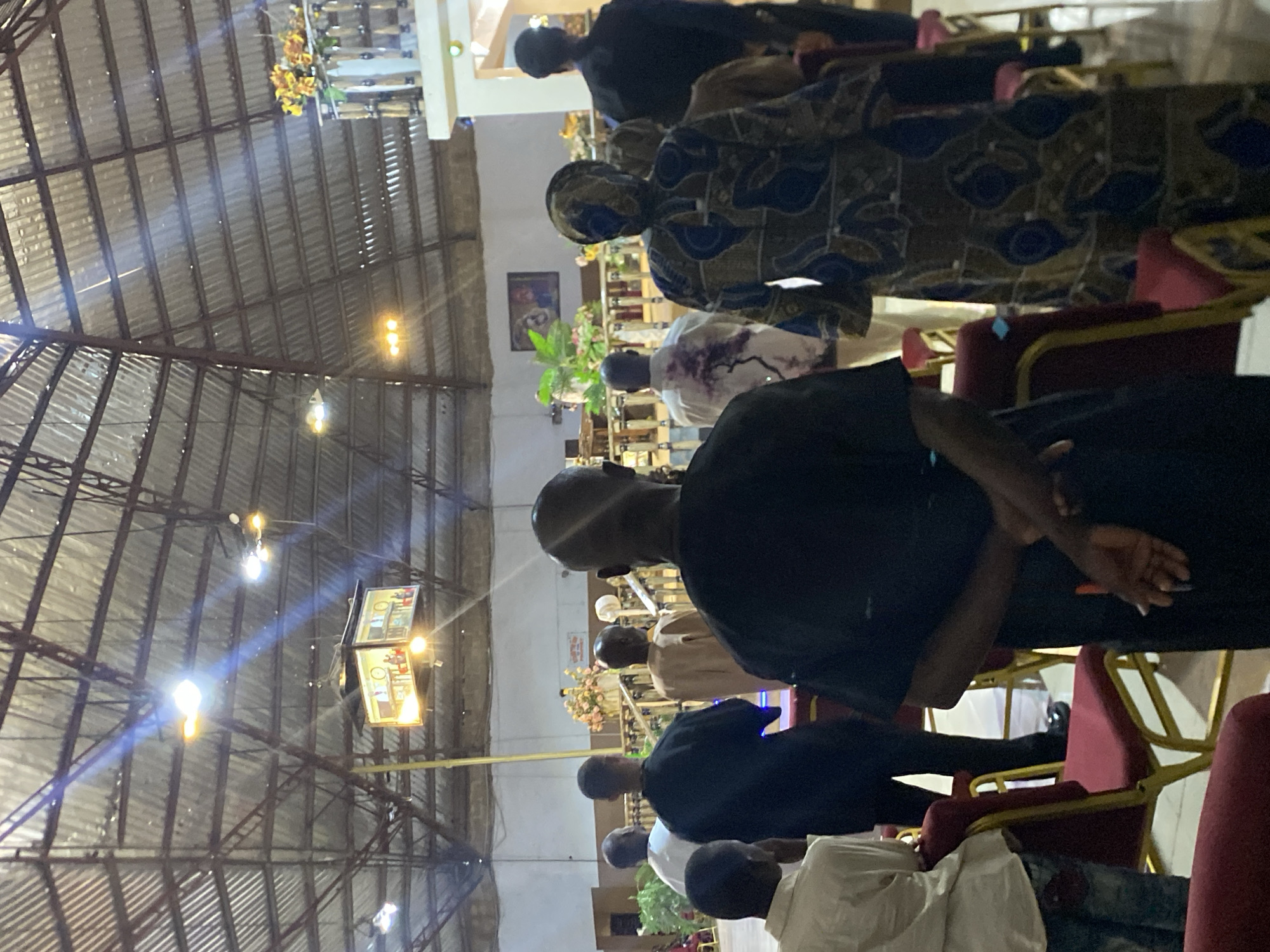 The image shows an indoor gathering of people in what appears to be a church setting, located on Rukuba Road, Nigeria. The building has a metal roof structure with visible beams and hanging lights. The attendees are seated in rows of red cushioned chairs. Several individuals are visible in the foreground and background, dressed in various clothing, including traditional African attire and Western-style clothing. Some appear to be praying.