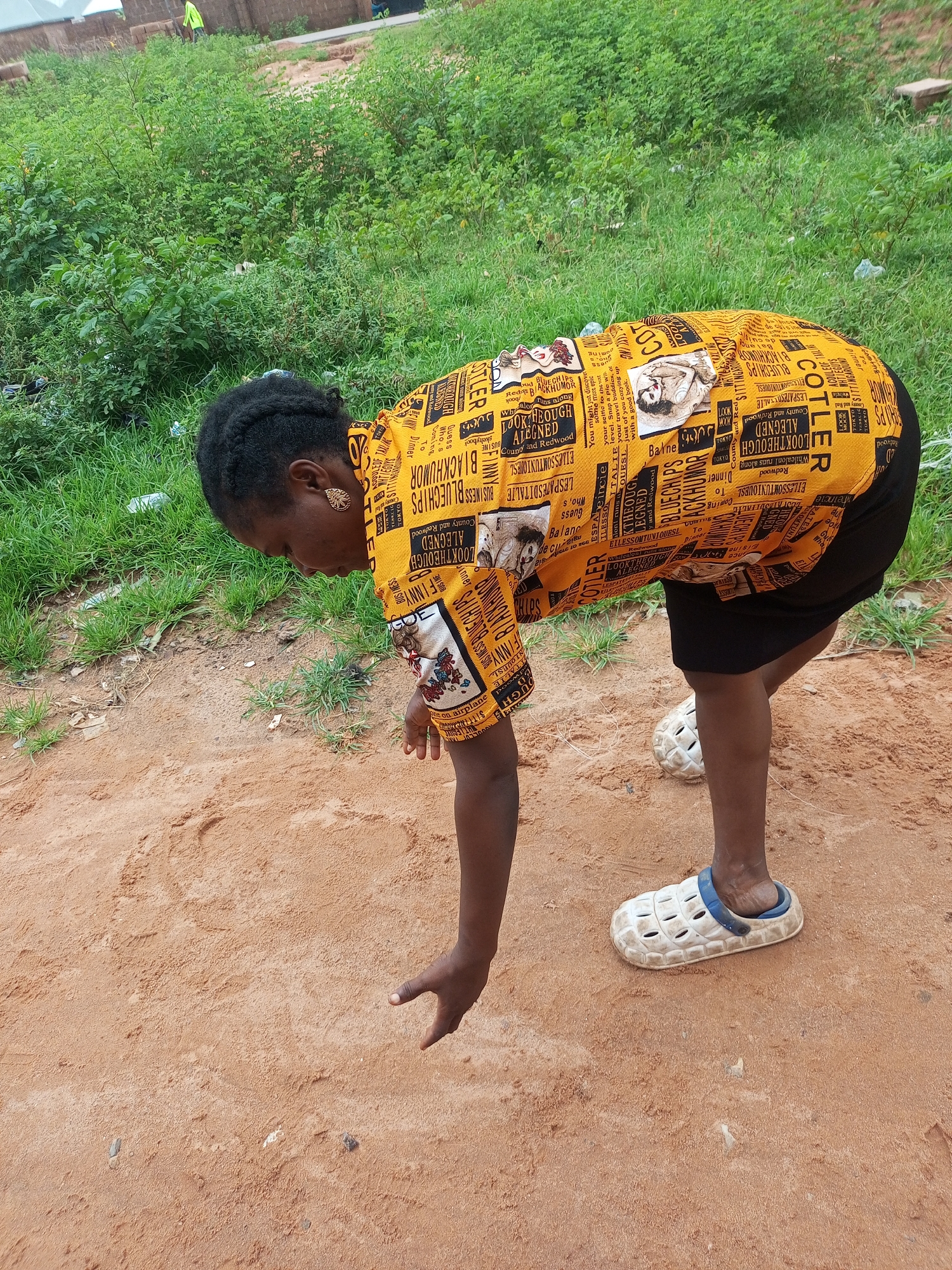 A woman, appearing to be of Nigerian descent, is captured in a medium shot, bending over and reaching her right hand towards the sandy ground. She wears a vibrant yellow, short-sleeved shirt adorned with a newspaper print pattern, featuring text and images. The shirt is tucked into a black skirt. Her footwear consists of white clogs with blue straps. Her hair is styled in braids, and she is wearing gold earrings. The environment is outdoors, with lush green vegetation and grass in the background. The ground beneath her is composed of reddish-brown sand, with some scattered small stones and debris. The scene suggests an outdoor setting in a non-urban area, possibly a rural or semi-urban location within Tempure, Nigeria.