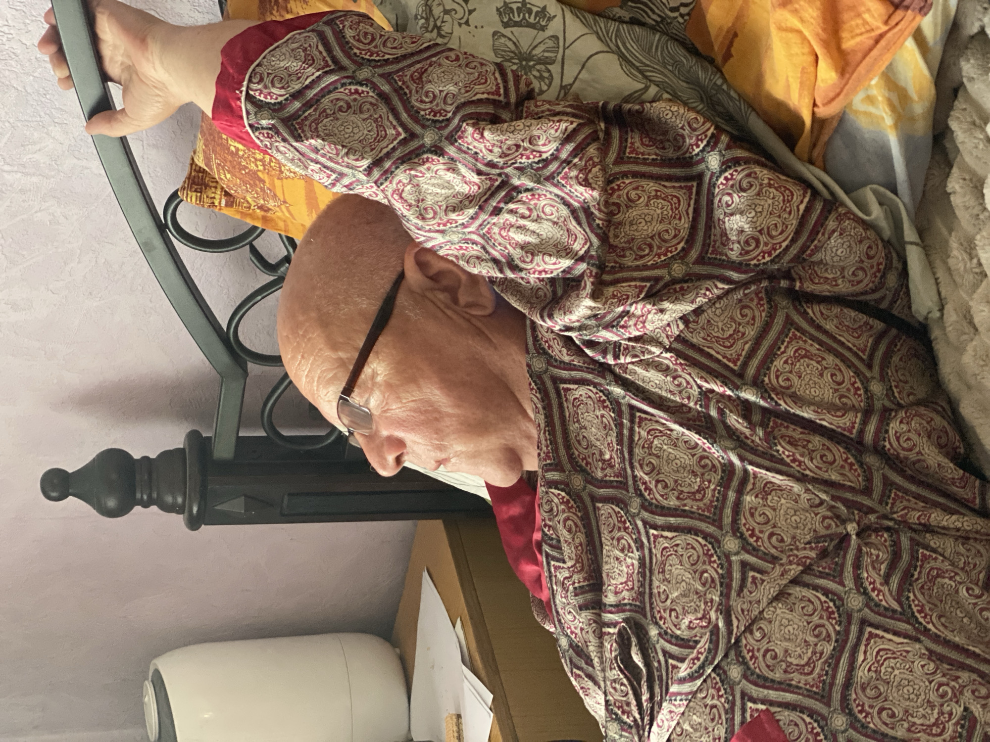 An elderly bald man wearing glasses is depicted lying in bed in a domestic setting, likely a bedroom in Novokuznetsk, Russia. He is positioned on his side, seemingly resting or sleeping. His right arm is extended upwards, grasping the dark metal headboard of the bed.
The man is dressed in a long-sleeved garment featuring an intricate pattern of red, beige, and dark grey elements, possibly a paisley or damask design, over what appears to be a plain red shirt or undershirt. Numerous pillows and blankets surround him, adding to the cozy atmosphere. Visible bedding includes patterned fabrics with designs such as butterflies, crowns, and feathers in muted tones, as well as solid colored textiles in orange and beige, and a fluffy, light-colored blanket.
Next to the bed, a wooden bedside table is partially visible, holding a white, cylindrical object that resembles a humidifier or air purifier, along with a stack of papers. The wall behind the bed is a light, textured color. The image shows no specific time of day or weather conditions. There is no visible text in the media file. The man's expression is neutral, suggesting a state of calm or slumber.