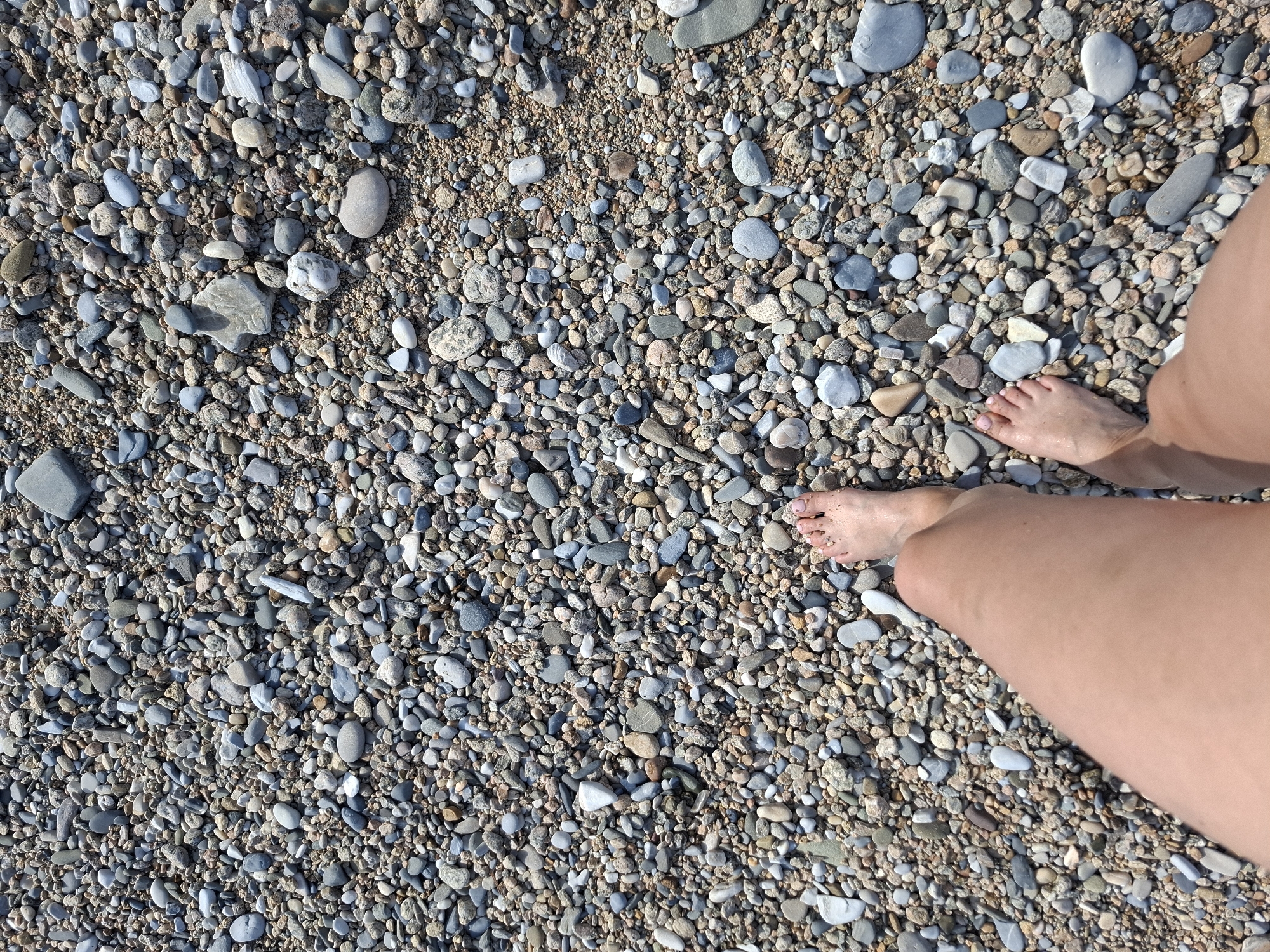 The image shows a person's legs and feet standing on a beach covered in pebbles. The pebbles vary in size and color, with shades of grey, brown, and white. The sun is shining, casting shadows on the pebbles. The water is not visible in the frame. The location is identified as Bugul'deika, Russia, suggesting this is a natural, outdoor scene.