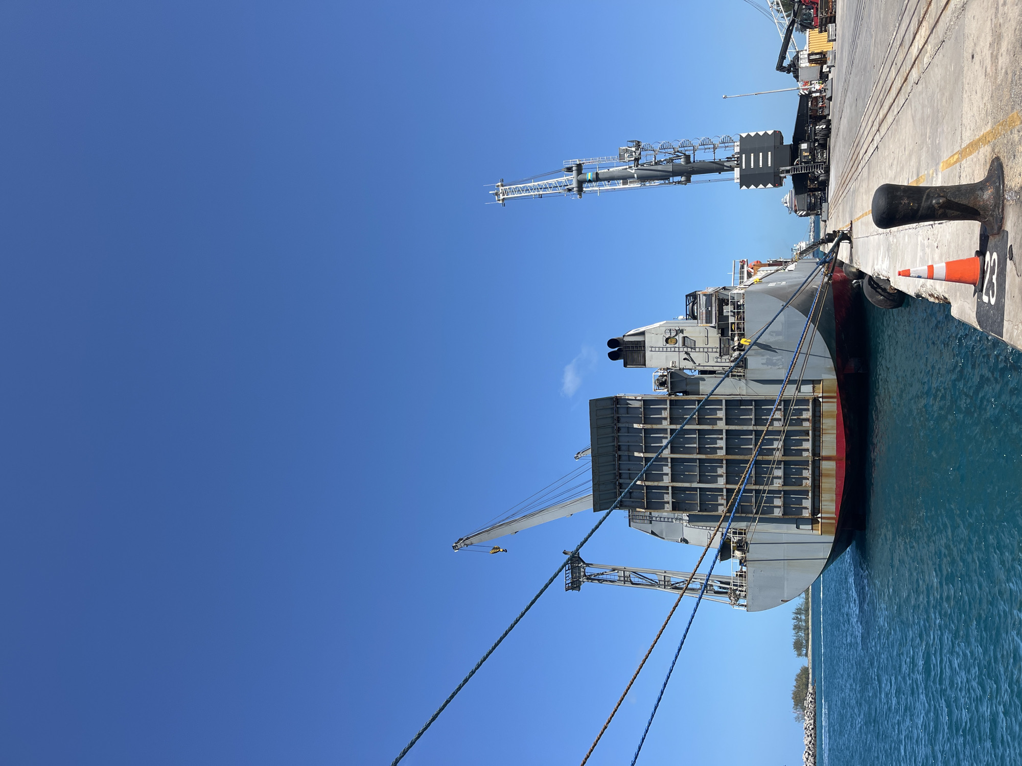 A container ship is docked at a pier on a sunny day. The ship is gray and red, with stacked containers visible on its deck. Mooring lines connect the ship to bollards on the concrete pier. A small orange and white traffic cone sits near a bollard marked with the number "23". Cranes are positioned alongside the ship on the pier. The sky is clear and blue. Water fills the space between the ship and a distant breakwater. The location is Unknown, Unknown.