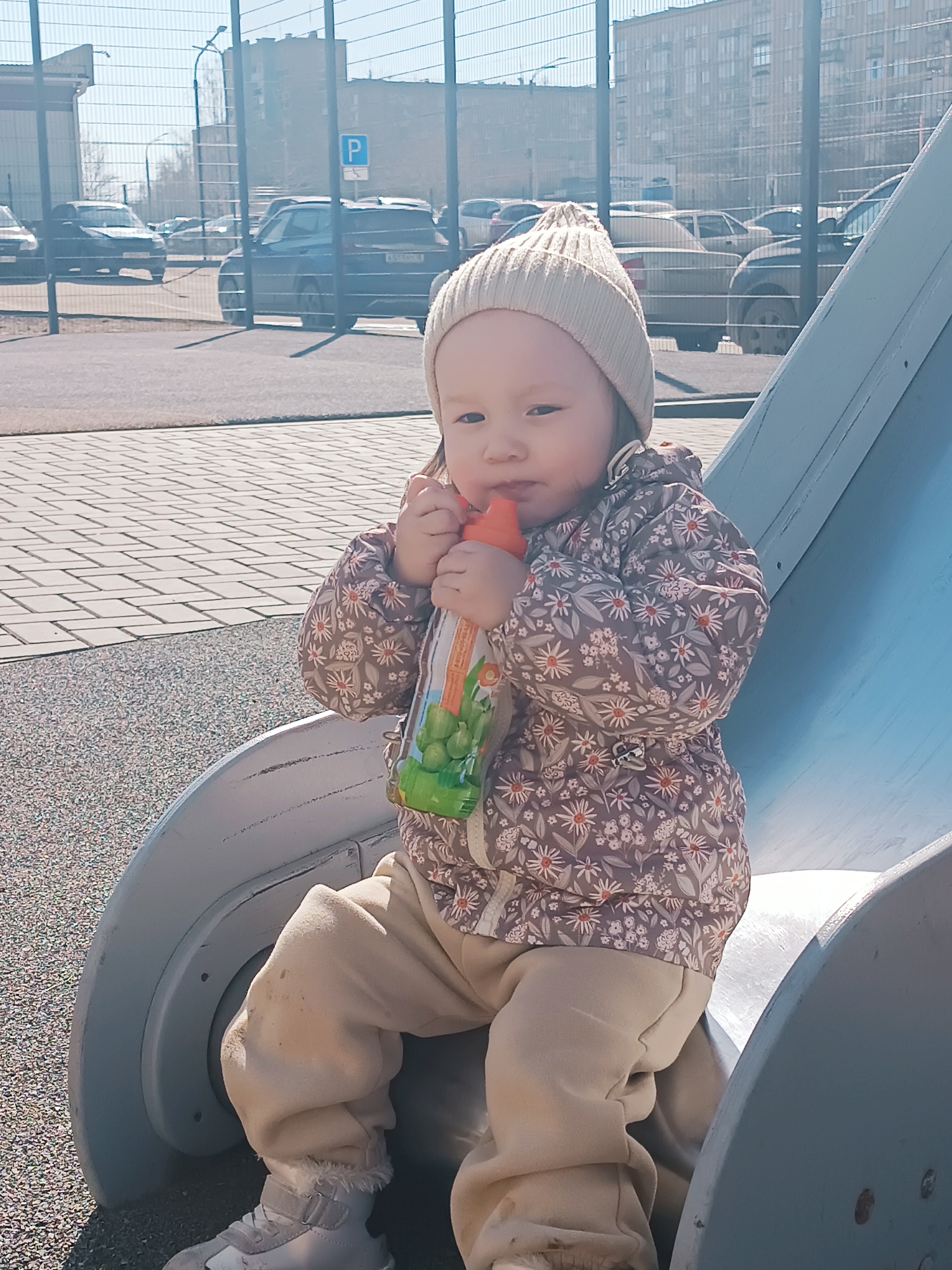 A young child in a beige hat, floral jacket, beige pants, and light-colored sneakers sits on a silver and blue slide outdoors. The child holds a bottle with green and orange packaging to their mouth. In the background, there is a parking lot with several cars, a fence, and buildings. A "P" parking sign is visible. The location is Izhevsk, Russia. The ground surface appears to be paving stones and a granular material, possibly playground surfacing. The day is sunny.