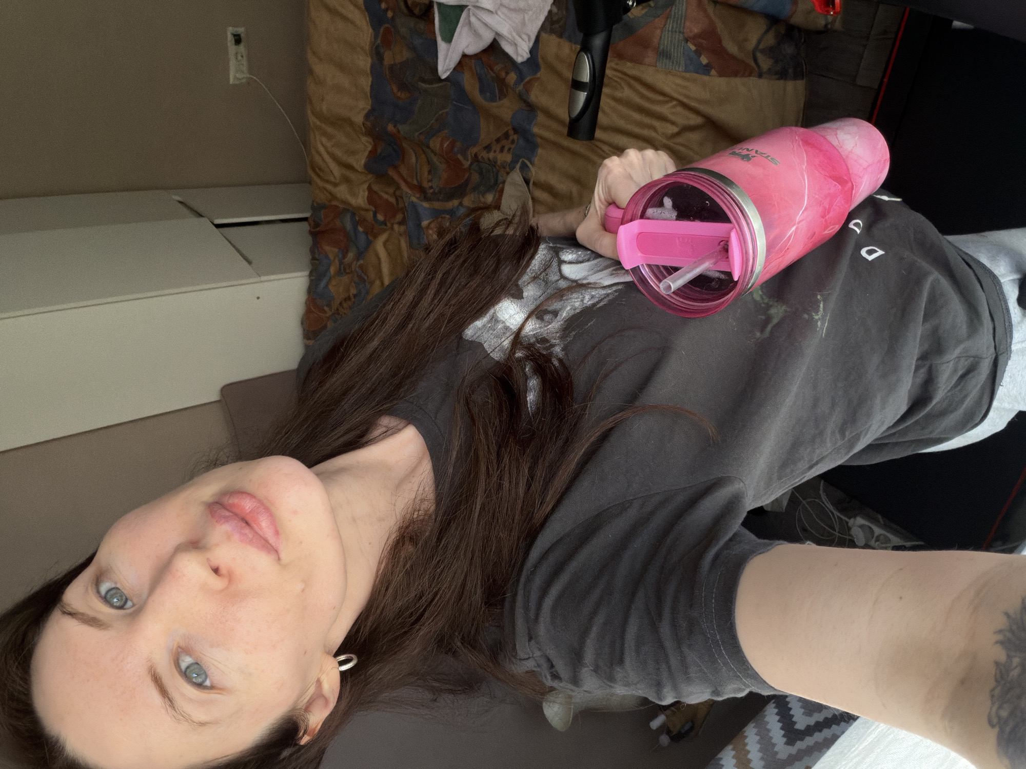 A person with light skin and blue eyes faces the camera, holding a pink Stanley brand tumbler with a lid and straw in their right hand. The person has long brown hair and is wearing a gray T-shirt. The background includes a bed with a patterned comforter, a white dresser, a charging cable, and an object that is most likely a paintball gun on the bed. The setting appears to be an indoor space. There is no information in the image to establish its location in Perm, Russia.