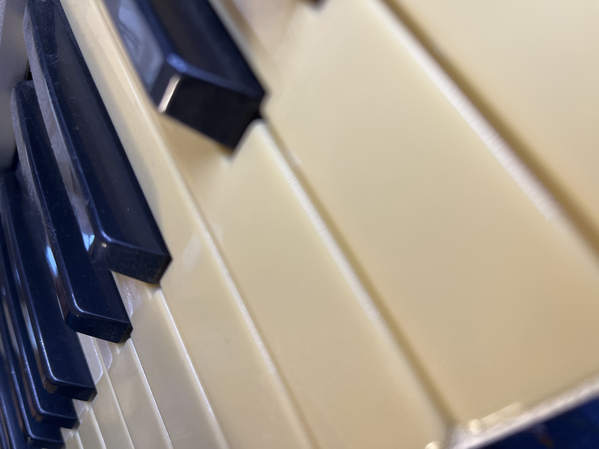 A close-up, angled shot focuses on a section of piano keys. The keys are made of a light yellow material, with the black keys positioned above them. The image is crisp, capturing the texture and slight wear on the keys. The background is a blur of muted colors, suggesting the keys are part of a larger piano. The scene does not depict any specific location in Krasnyye Barrikady, Russia, nor does it include any people or actions.