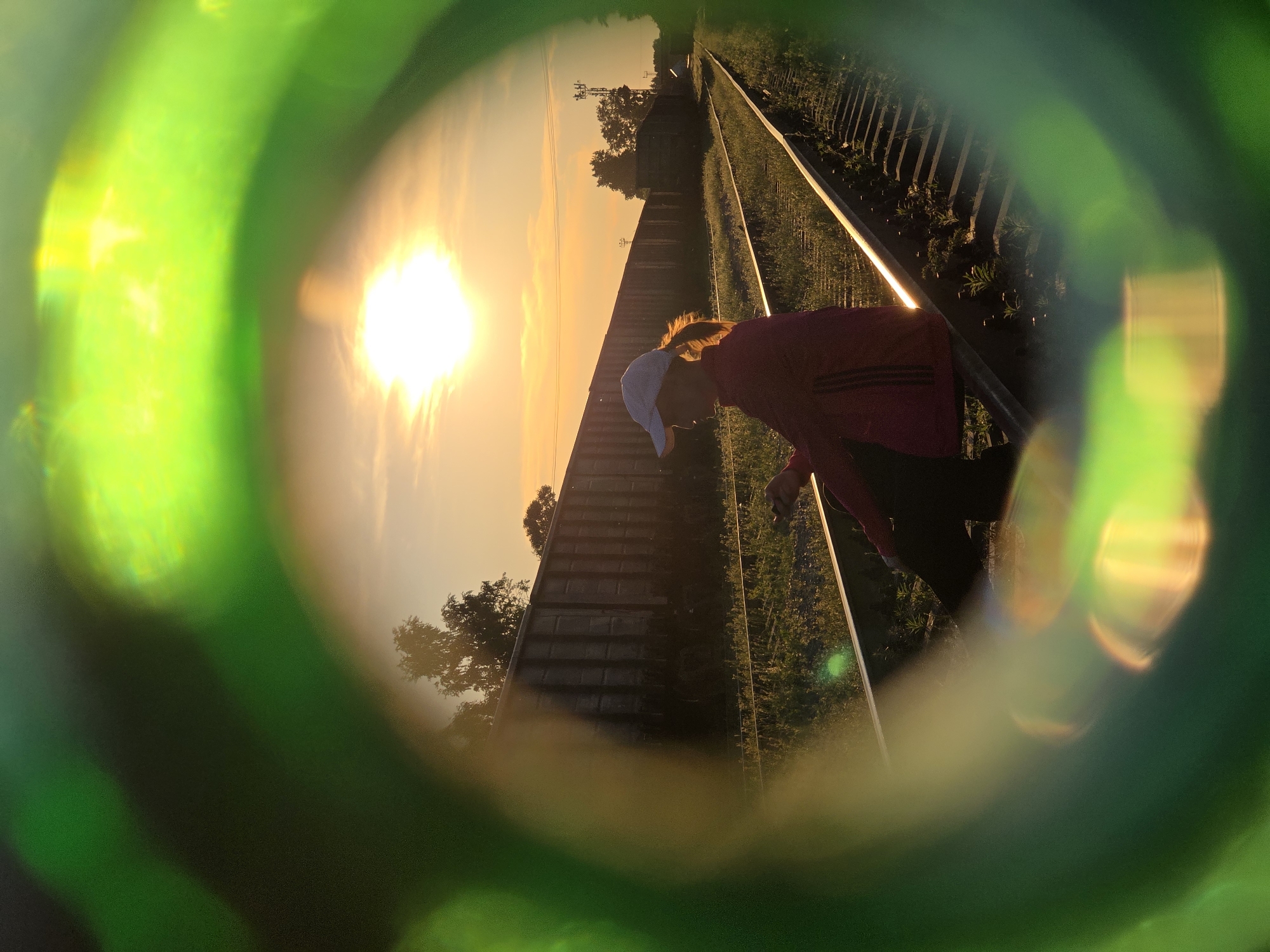 An individual is seated on railway tracks at a location identified as Chaikovsky, Russia. The person is wearing a pink jacket, dark pants, and a white baseball cap. The setting appears to be outdoors during sunset, with the sun visible in the sky. The railway tracks extend into the distance, lined with freight train cars on one side. Green foliage grows along the sides of the tracks. The image is framed by a blurry green element, suggestive of looking through a glass object. The overall lighting indicates a warm, late afternoon or early evening atmosphere.