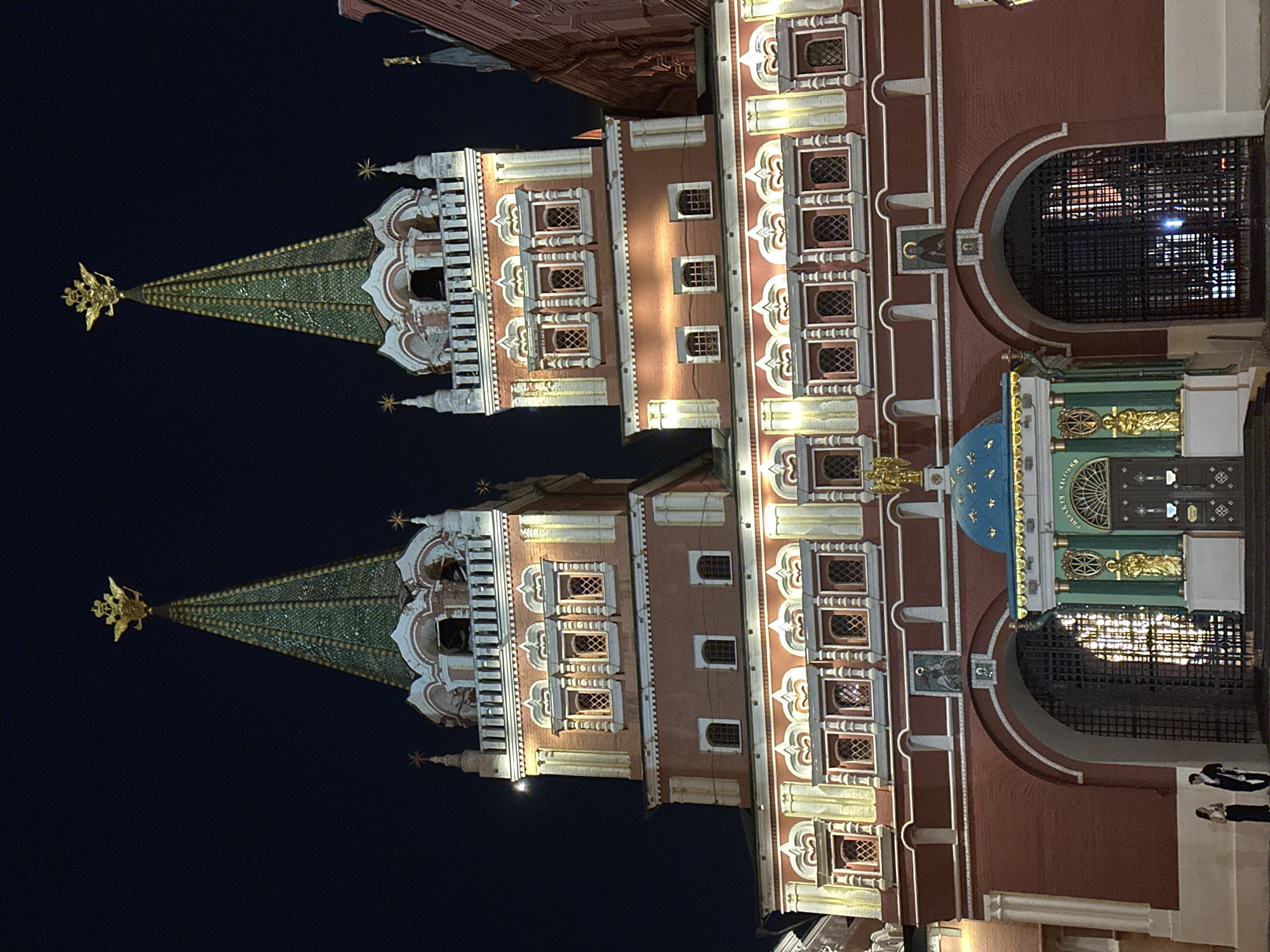 The image, when viewed upright (rotated 90 degrees clockwise from its current orientation), displays two perfectly identical, ornate historical towers, illuminated at night. Each tower features a tall, conical spire covered in green, scale-like tiles, culminating in a golden double-headed eagle at its apex. The main structure is a reddish-brown building adorned with numerous white decorative architectural elements, including cornices, window frames, and pilasters. Multiple rectangular windows with intricate grates are visible across the facade.
The rightmost tower (when viewed upright) includes a large, semicircular arched entrance or passage, secured by a dark metal grate. Below this arch, a richly decorated panel features a small blue dome, green sections, and golden sculptural elements, including figures. Indistinct Cyrillic text is faintly visible on this decorated section. The entire building is illuminated by artificial lights, casting warm glows against the dark night sky. In the lower right corner, two indistinct human figures are standing near the base of the building. The architectural style, including the double-headed eagle, strongly indicates a location within Moscow, Russia, likely a part of the Kremlin complex.