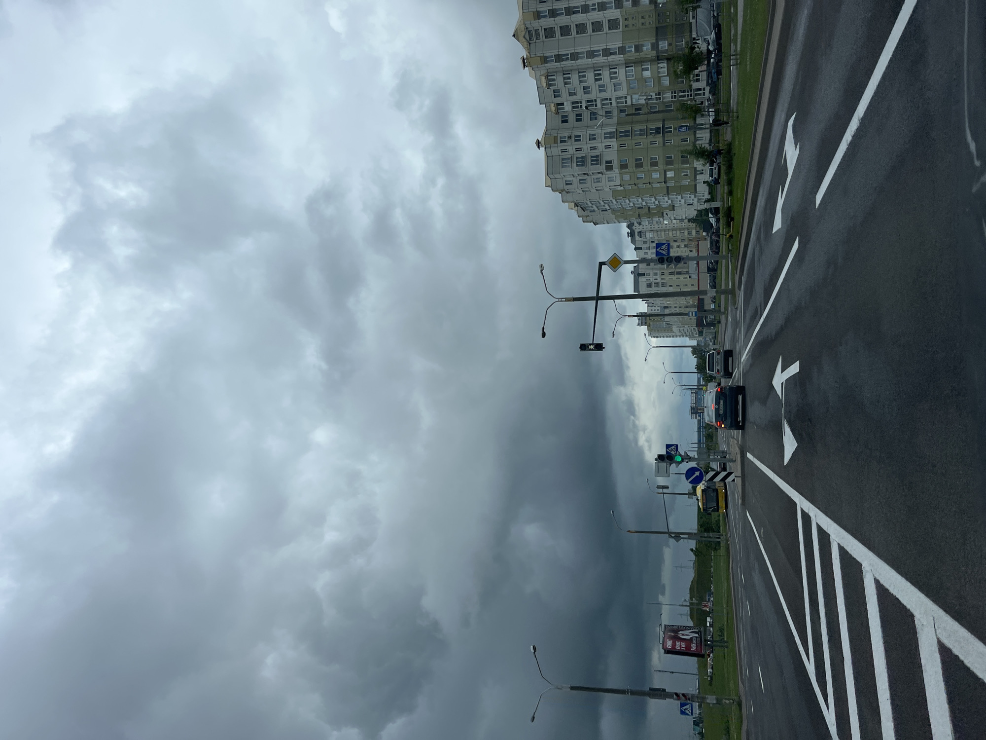 This is a wide, slightly low-angle, full shot of a road in Minsk, Belarus, under a heavily overcast sky. The perspective is from within a vehicle, looking forward down a multi-lane asphalt road. The road is wet, indicating recent rain, and marked with white lines, including a pedestrian crossing in the foreground on the left and directional arrows on the right.
In the mid-ground, a yellow bus is turning left, and a dark-colored sedan is stopped at a traffic light. The traffic light is green. Several tall, modern apartment buildings with many windows line the right side of the road, their facades a mix of grey, white, and green. On the left side of the road, there are streetlights, traffic signs, and a billboard. The sky is dominated by dark, dramatic storm clouds, with a lighter patch of sky visible in the distance on the right. The overall atmosphere is somber and expectant of more rain. No individuals are clearly visible or identifiable.
