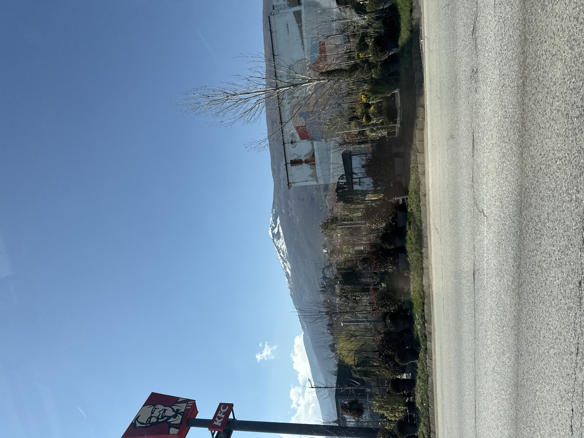 The image depicts a street scene in Sofia, Bulgaria, with a prominent KFC sign in the foreground. A road stretches horizontally across the lower part of the frame. Adjacent to the road, there's vegetation and buildings. In the distance, a mountain range is visible. The sky is clear blue with some scattered clouds. A large, weathered building with fading murals or paintings can be seen beside the road. The overall impression is of a landscape blending urban and natural elements.