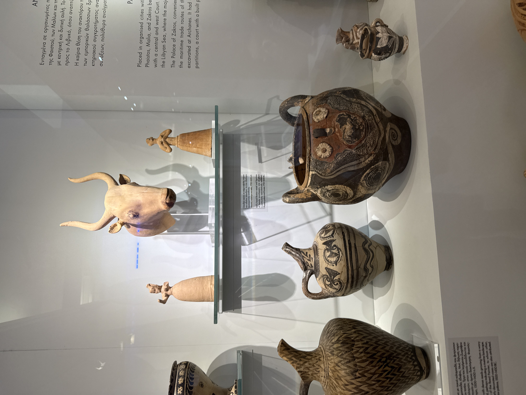 The image shows a display of ancient Minoan artifacts in a museum. The objects are housed in a glass-fronted display case with white walls and shelves. On a top shelf, there is a wooden sculpture of a bull's head and two small clay figurines of women. Below this, on a lower shelf, are several large pottery vessels. The closest vessel on the left is a tall, dark-colored vase with a pattern of intersecting lines. To its right is a lighter-colored jug with black painted decorations. In the center of the lower shelf is a large, bulbous pot with elaborate black and red painted designs, including circular motifs and figures. To the right of this, a smaller, dark-colored jug with white painted decorations is visible. A text panel is partially visible on the right side of the display case, and another text panel with Greek and English descriptions is placed on the lower shelf in front of the pottery. The overall setting suggests an archaeological exhibition. The text panel indicates that the artifacts are from the city of Heraklion, Greece, and are associated with organized cities, palaces, and maritime trade routes.