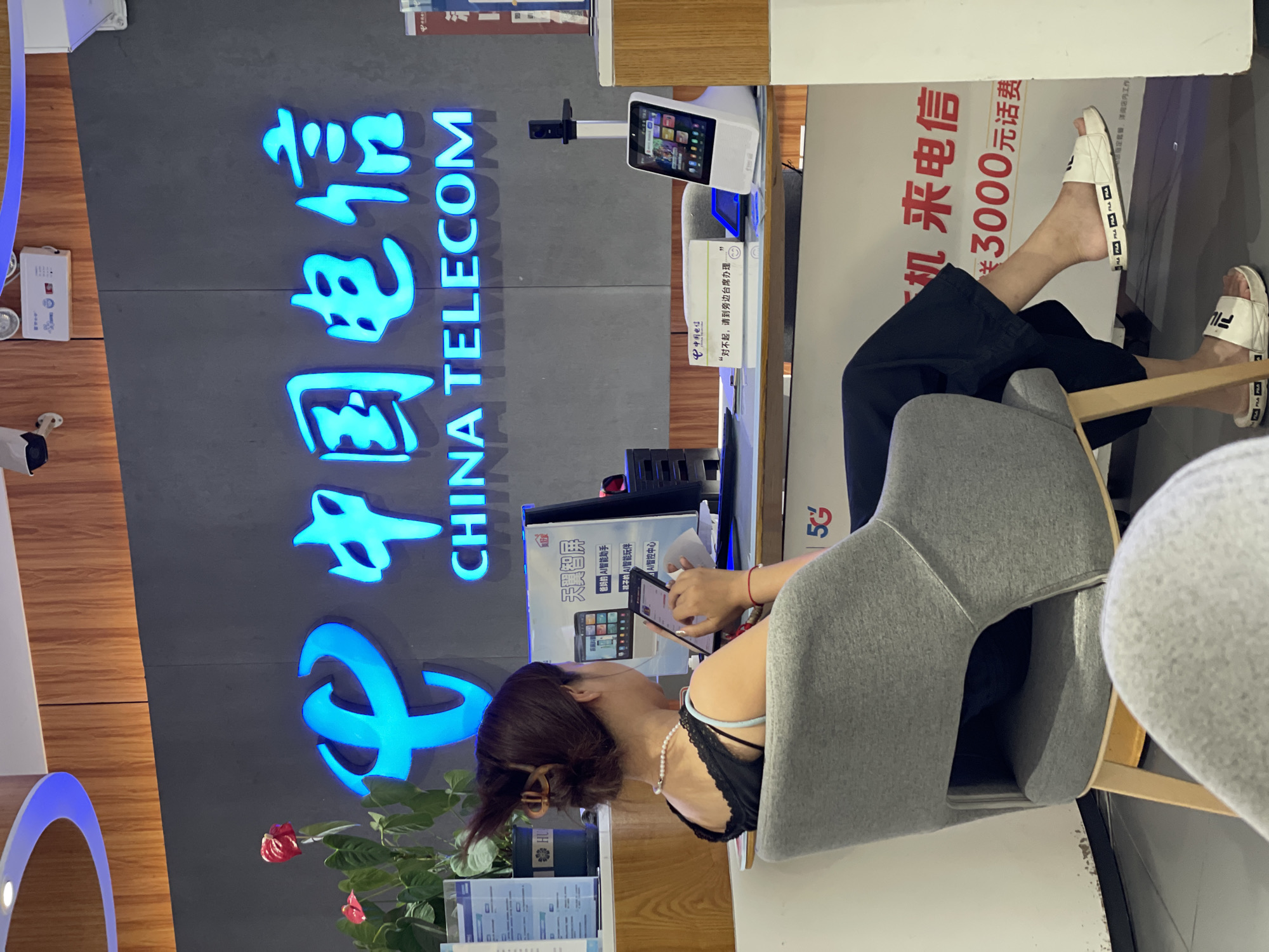 The image shows the interior of a China Telecom store in Nan'an District, China. A woman is seated in a grey armchair, facing a counter and using her smartphone. Behind the counter, a large, illuminated "CHINA TELECOM" logo is visible on a dark grey wall. Various electronic devices and promotional materials are displayed on the counter, including a tablet and a smart display. A potted plant with red flowers is situated to the left of the logo. The overall environment suggests a modern retail space designed for telecommunications services.