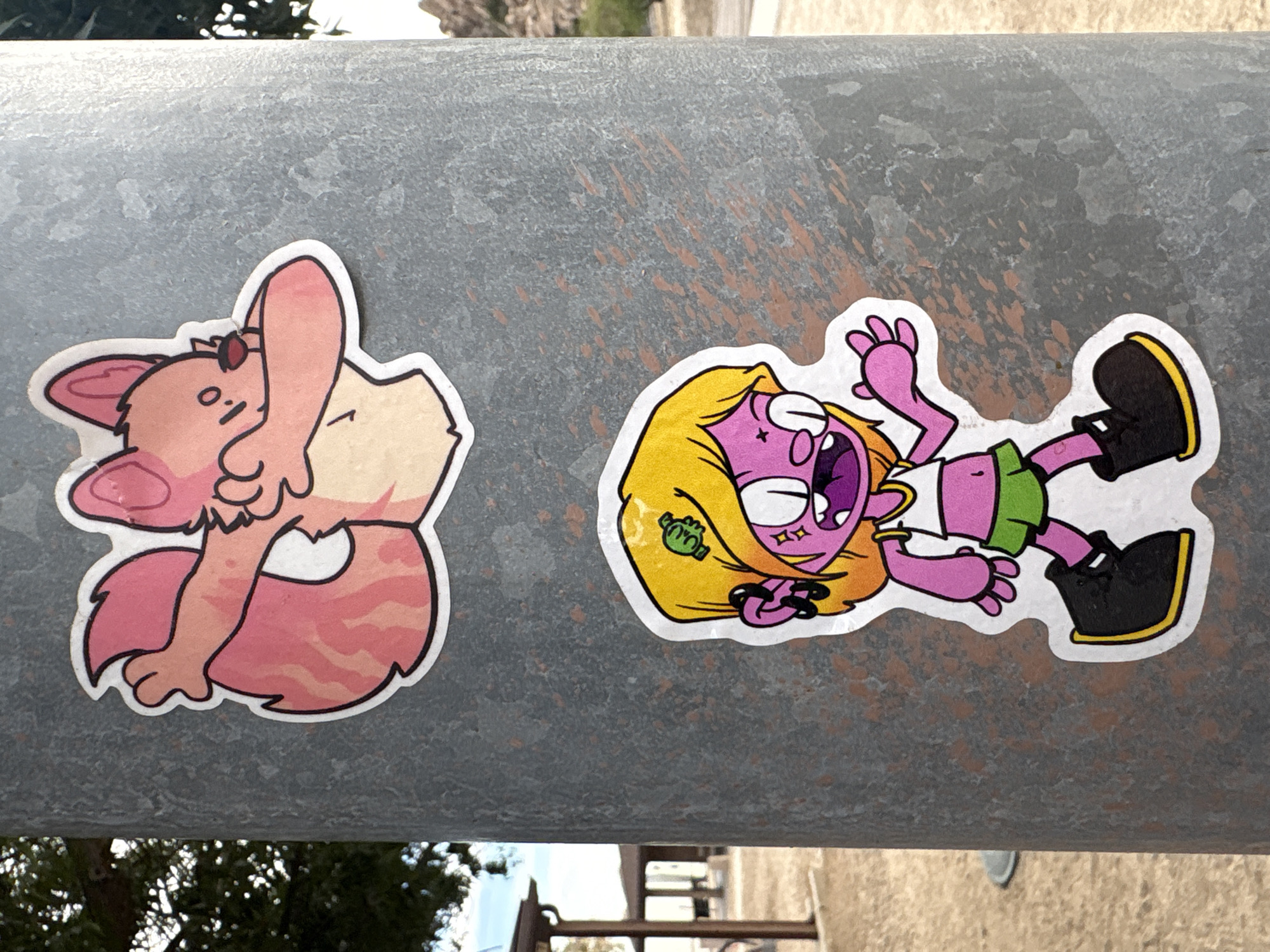 Two stickers are affixed to a gray metal pole in Dragoon, Arizona. The top sticker features a pink fox-like character with large ears and a fluffy tail. The character is in a pose with one arm bent and the other raised, covering its face. The bottom sticker depicts a cartoonish character with bright yellow hair, purple skin, and a wide, open mouth. This character wears a white crop top, a green skirt, and black boots with yellow trim. The background appears to be an outdoor setting with trees and a building visible in the distance.