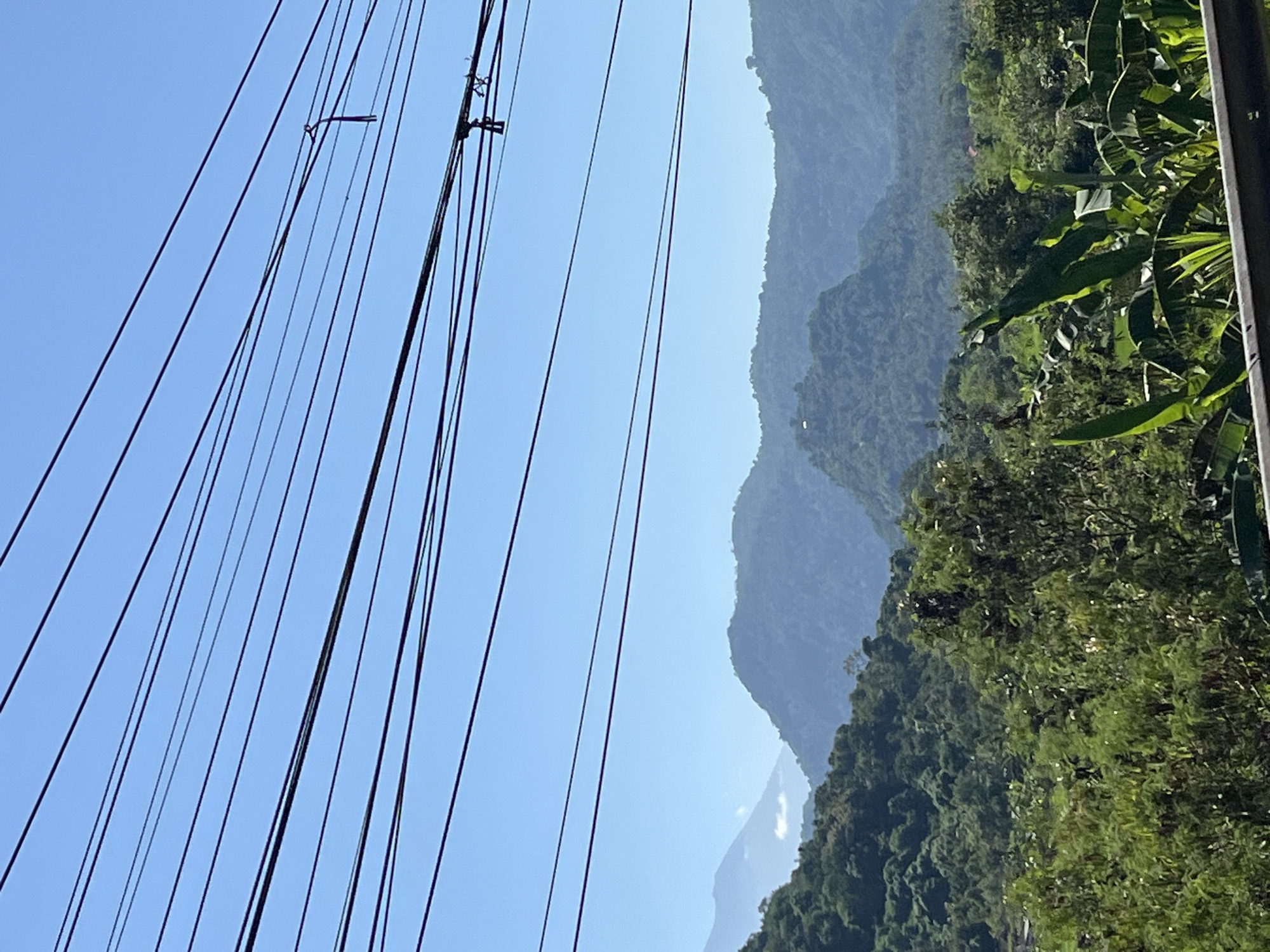The image captures a scenic view of mountains and lush greenery, typical of the landscape in Dawan, Indonesia. Power lines are prominently strung across the foreground against a clear blue sky. In the background, rolling hills are covered in dense vegetation, with some appearing to be cultivated or have small structures on them. The distant mountains fade into a hazy blue, suggesting a broad and expansive vista. The lower part of the image shows more detailed foliage, including large leaves that could be from banana plants, indicating a tropical environment. There are no people visible in the frame. The scene is static and peaceful, showcasing the natural beauty of the region.