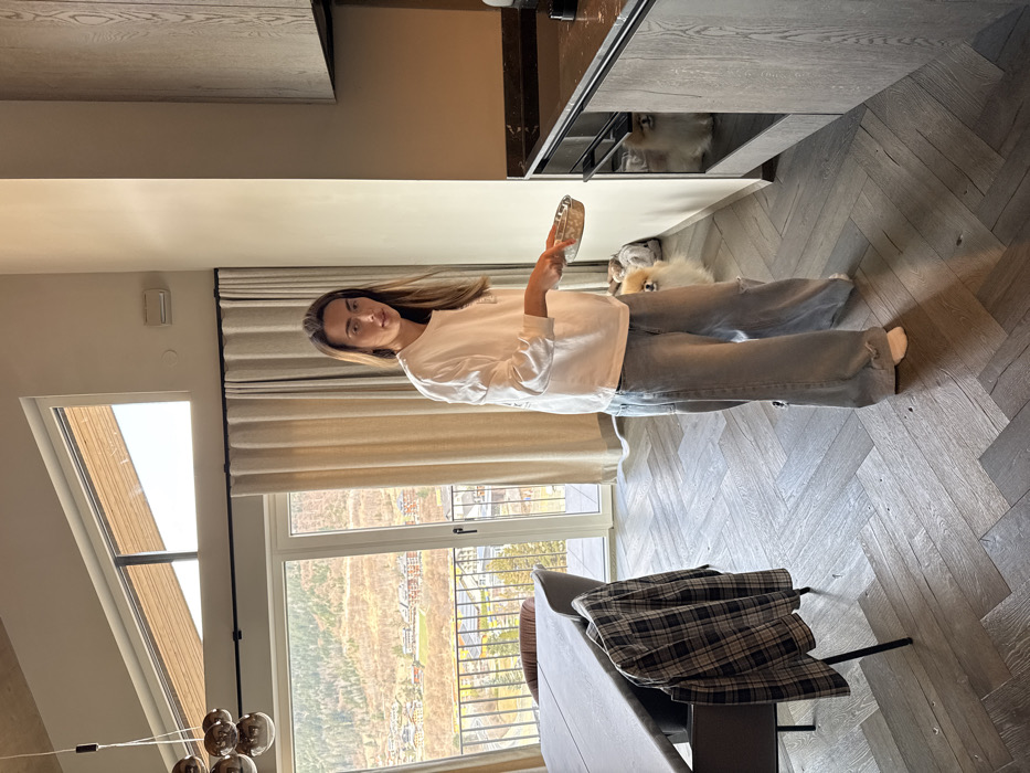 The image captures an indoor scene within a modern residence in Schladming, Austria, featuring a panoramic view of mountainous terrain and a valley town. A Caucasian female, with long brown hair parted in the middle, stands in the center-right of the frame. She wears a white long-sleeved t-shirt and light blue distressed wide-leg jeans, looking directly towards the camera. She holds a small, silver-colored bowl in her extended right hand, positioned towards the kitchen area. Two small, fluffy, light-colored dogs, identified as Pomeranians, are present; one stands on the dark herringbone wooden floor behind the woman's left leg, while the other is visible within an open lower shelf of the dark wood kitchen cabinetry to her right. The kitchen area on the right features dark wooden cabinets with a horizontal grain and a dark countertop. To the left, a dining area includes a dark grey rectangular table with a matching upholstered chair, over which a black and grey plaid shirt is draped. Large glass sliding doors and a high, triangular window in the background provide extensive natural light and a clear view of a mountainous landscape with scattered buildings and green/brown vegetation. A black metal balcony railing is visible immediately outside the glass doors. A modern chandelier with multiple spherical glass globes hangs from the light-colored, slanted ceiling in the upper left.