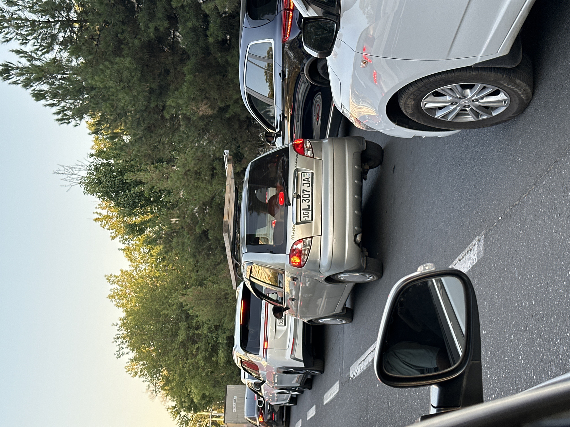 The image depicts a traffic jam on a city street, likely in Tashkent, Uzbekistan, given the license plate format visible on the silver Daewoo Matiz. The time of day appears to be late afternoon or early evening, with the sun casting warm light on the scene and trees lining the road.
In the foreground, the side mirror of a white vehicle is visible, reflecting the interior and a person wearing a light green shirt. The road is paved and marked with white lane lines.
Several cars are stalled in traffic, moving from left to right. The most prominent vehicles are:
- A silver Daewoo Matiz, with license plate "10 L 307 JA" (Uzbekistan plates). It is carrying what appears to be a ladder or long planks on its roof.
- A white SUV, possibly a Foton, with a modern design and illuminated taillights. Its license plate begins with "01".
- A dark blue SUV or crossover, with tinted windows and distinctive taillight design.
- Another white vehicle is partially visible in the bottom right corner, showing its front wheel and part of its bodywork.
- Further back, a black car is visible.
- A white van with signage "502 ADA" is seen in the background on the left.
The background consists of a dense line of green trees and a clear sky. The overall atmosphere suggests a typical urban commute, possibly during rush hour.