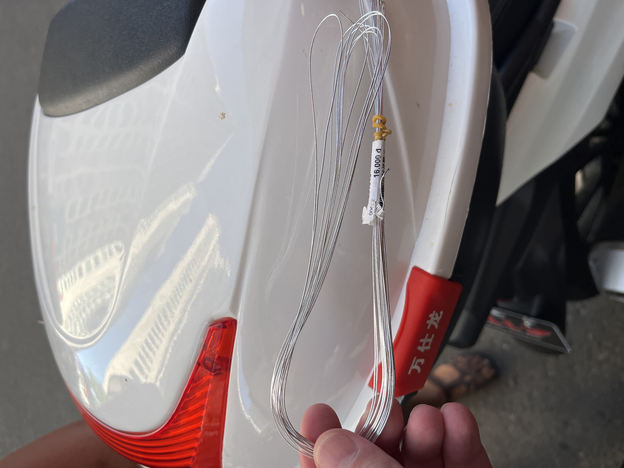 A hand holds a bundle of silver-colored wires secured with a rubber band and a label marked "16.000 đ" against the backdrop of a white and red motorcycle in Bac Nha Trang Ward, Vietnam. A person's sandaled foot is visible in the background to the right of the motorcycle.