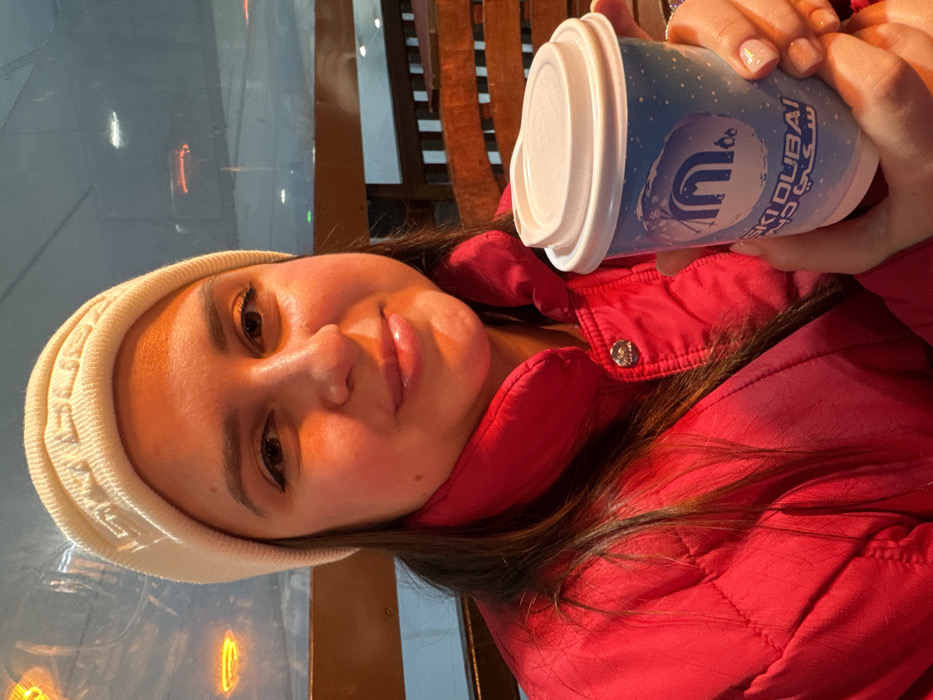 A female subject is photographed from the chest up, positioned indoors at Ski Dubai in the Mall of the Emirates, Dubai, United Arab Emirates. She faces the camera with a slight smile. The subject wears a cream-colored knit beanie, embossed with text identified as "SKI DUBAI", and a bright pink quilted jacket with a high collar. Her dark hair is visible below the beanie. She holds a light blue paper cup with a white plastic lid in both hands. The cup features a white speckled design, a stylized 'M' logo, and "SKI DUBAI" text in English and Arabic. The background includes reflections of orange lights on a large glass panel. Behind the glass, a pale blue-white environment consistent with an indoor ski slope is discernible. A wooden railing is partially visible to the subject's right. Warm lighting illuminates the subject's face.