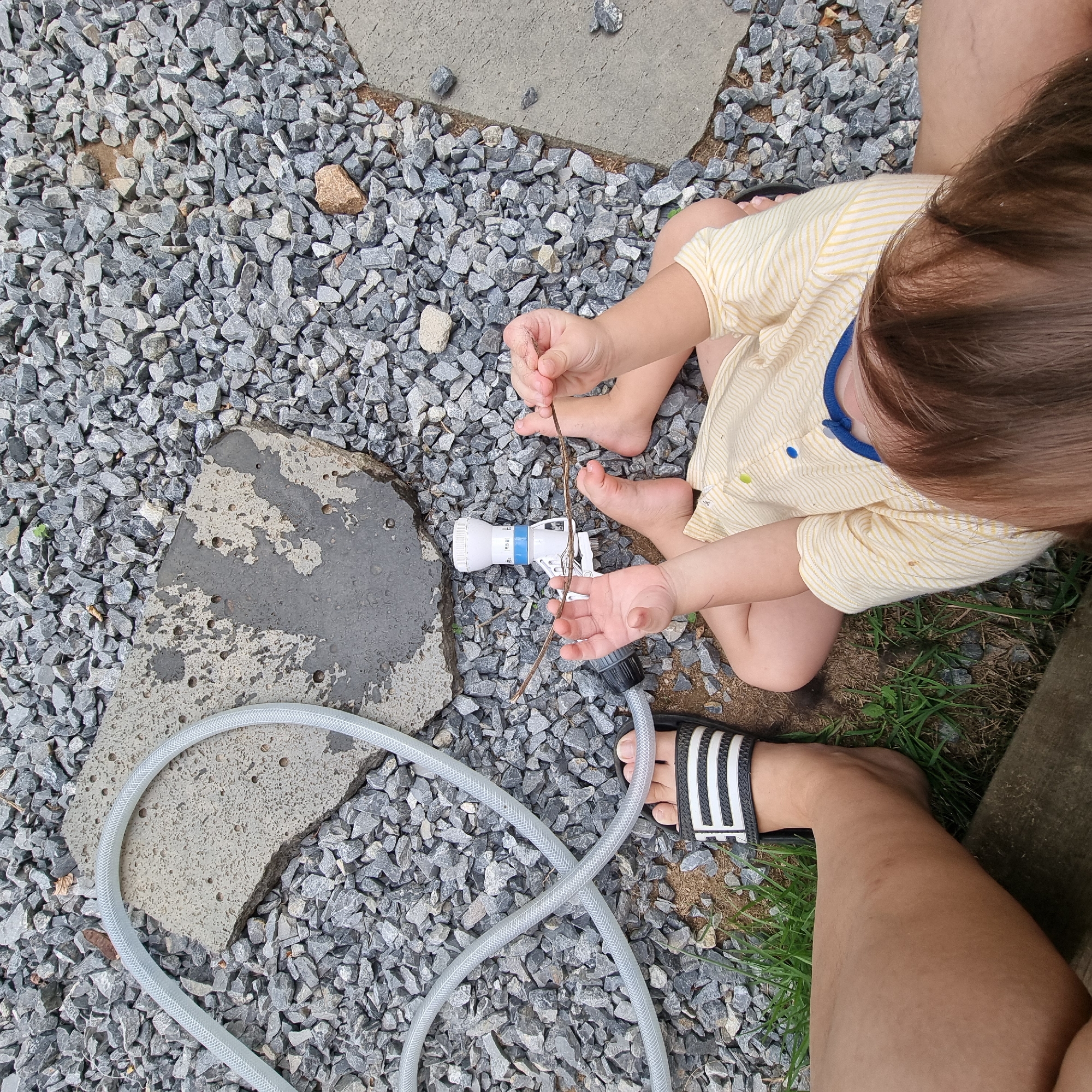 In Dunnae-myeon, South Korea, an aerial shot reveals a child sitting on a gravel surface, holding a thin stick. The child, wearing a yellow and white striped shirt, has their legs bent and feet bare except for one foot adorned with a black and white striped sandal. To the left of the child, an adult's leg, clad in a similar sandal, is visible, suggesting they are seated next to the child. A white and blue water sprinkler nozzle lies on the ground between the child's hands, and a translucent hose, likely connected to the sprinkler, snakes across the gravel. The environment is primarily composed of gray gravel with patches of dirt and sparse green grass, interspersed with two flat concrete slabs.