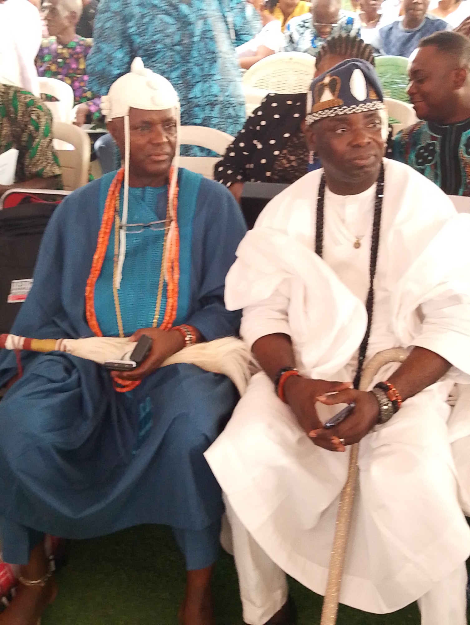 Two men in elaborate traditional attire are seated at what appears to be a community gathering or ceremony in Dushepe, Nigeria.
The man on the left wears a distinctive white cap, a flowing blue tunic with a lighter blue textured panel, and multiple strands of bright orange coral beads around his neck. Eyeglasses hang from one of the beaded necklaces. He holds a dark smartphone in his right hand and a red and yellow marked traditional staff or wand in his left, resting on his lap. His left ankle is adorned with a golden anklet, and his feet appear bare.
The man on the right is dressed in a white flowing tunic and a dark blue cap intricately embroidered with white and gold patterns. A long black beaded necklace hangs around his neck, and he wears various bracelets on both wrists, including orange and dark ones. He holds a dark smartphone in his left hand, while a golden, ornate staff or cane rests vertically between his legs.
Both men have composed expressions, looking slightly to the side. They are seated on white chairs, with other attendees in traditional and casual clothing visible in the blurred background. The ground is covered with green artificial turf or carpet. The lighting suggests it is daytime, possibly in an indoor or shaded outdoor setting. Their attire and staffs indicate they are likely respected figures or chiefs at the event. No discernible text is visible in the image.