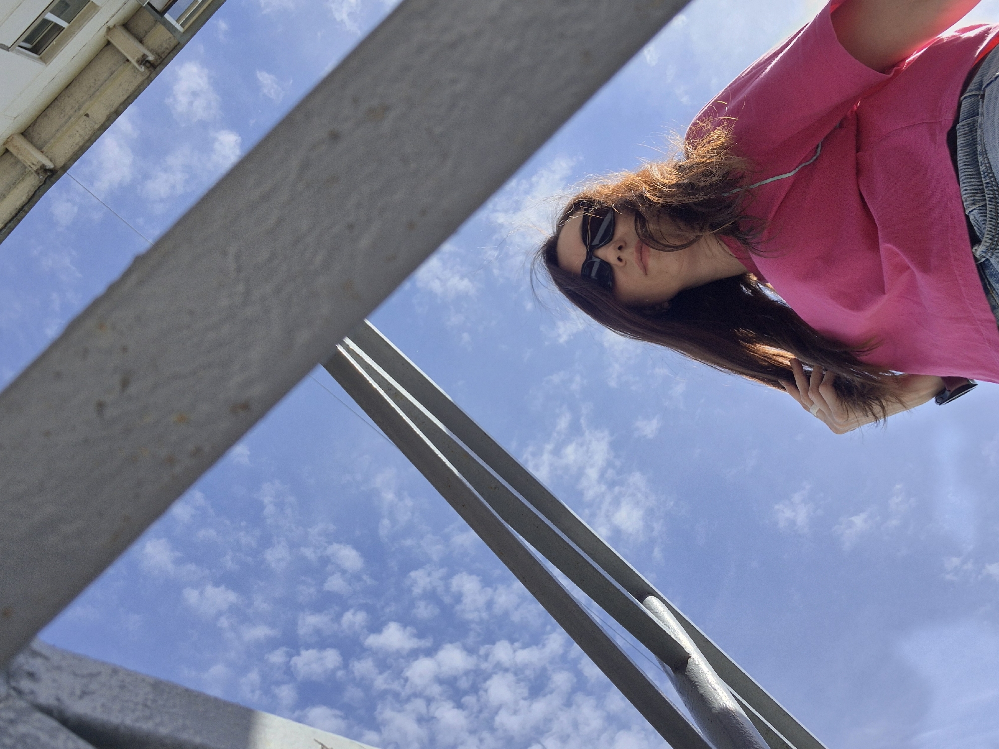 The image shows a low-angle view looking up through a metal structure towards a woman with sunglasses and a pink shirt, set against a backdrop of a blue sky with scattered clouds. Part of a building is visible in the top left corner of the frame. The location appears to be a city, but the specific details needed to identify the city or the building type are not available in this picture.