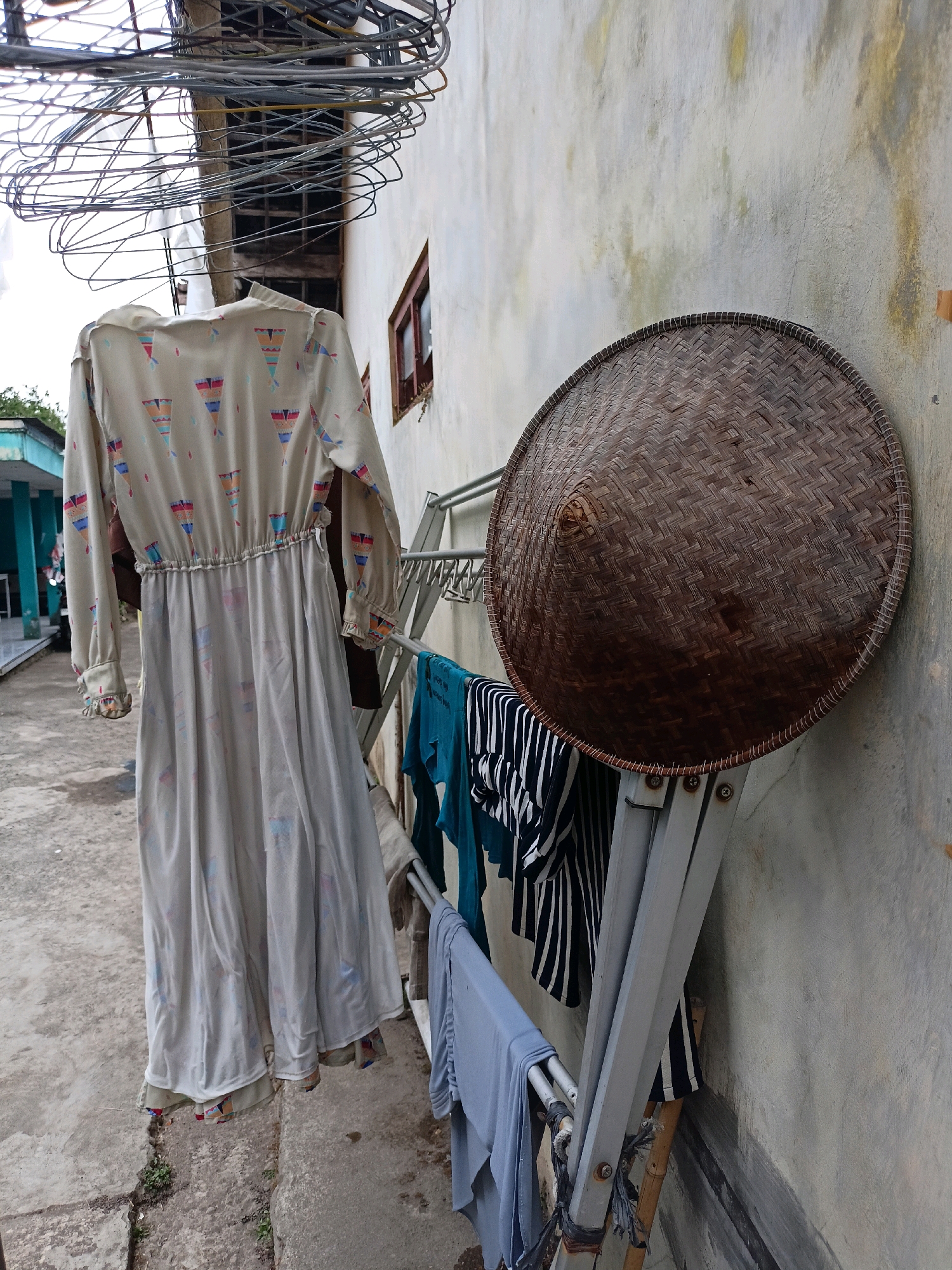 In Ngepanrejo, Indonesia, a light-colored dress with a patterned print is hanging from a clothesline. Nearby, a large conical woven hat is propped against a wall. Other clothes, including a striped shirt and blue fabric, are also drying on a metal rack. The scene appears to be an outdoor drying area next to a building with a rough, textured wall. Overhead, a tangle of wires and clothes hangers are visible. The ground is concrete, and some sparse vegetation is growing through cracks. The lighting suggests an overcast day.