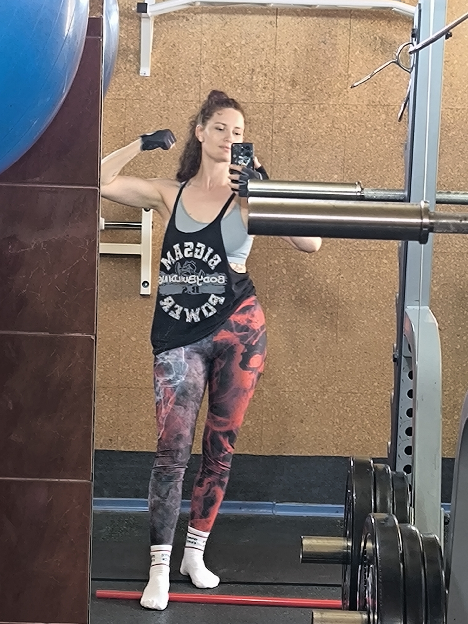 In a gym setting, a woman is captured in a mirror selfie. She is wearing a black tank top with a graphic print and grey undergarment, paired with patterned leggings and white socks. Her left arm is flexed, showcasing her bicep. The background reveals gym equipment, including a weight rack with plates, and a large blue exercise ball. The walls are covered in cork. The image appears to have been taken in a gym in Saint Petersburg, Russia.