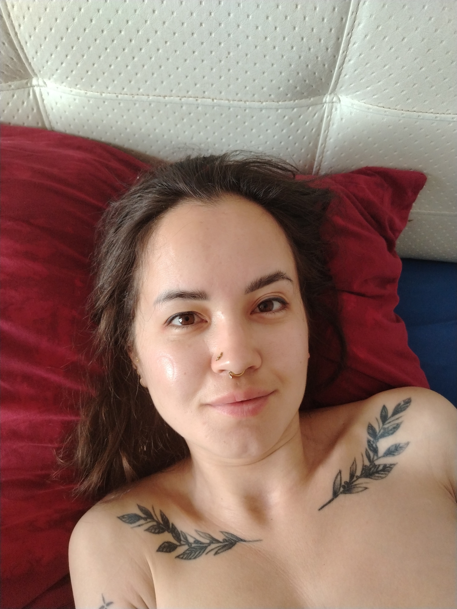 A woman lies on a bed with a red pillow. She has brown hair, brown eyes, and a nose ring. She has two leaf tattoos on her chest. The bed has a white headboard and a blue sheet. The location is Noviy, Russia.