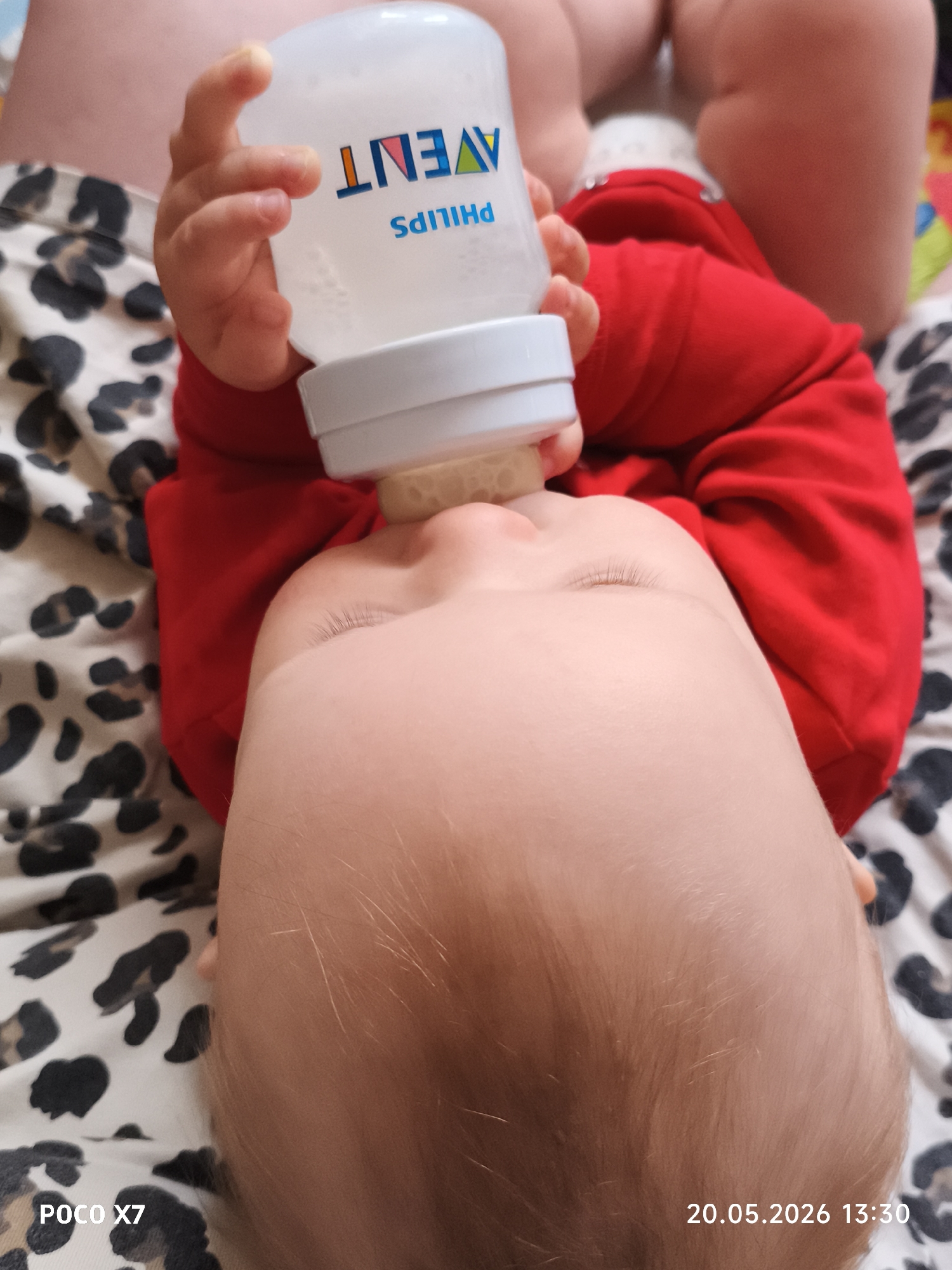A baby in a red shirt is lying on a leopard-print blanket and drinking from a Philips Avent bottle. The timestamp indicates the photo was taken on May 20, 2026, at 13:30. The location is Chaikovsky, Russia, based on the city in the query.