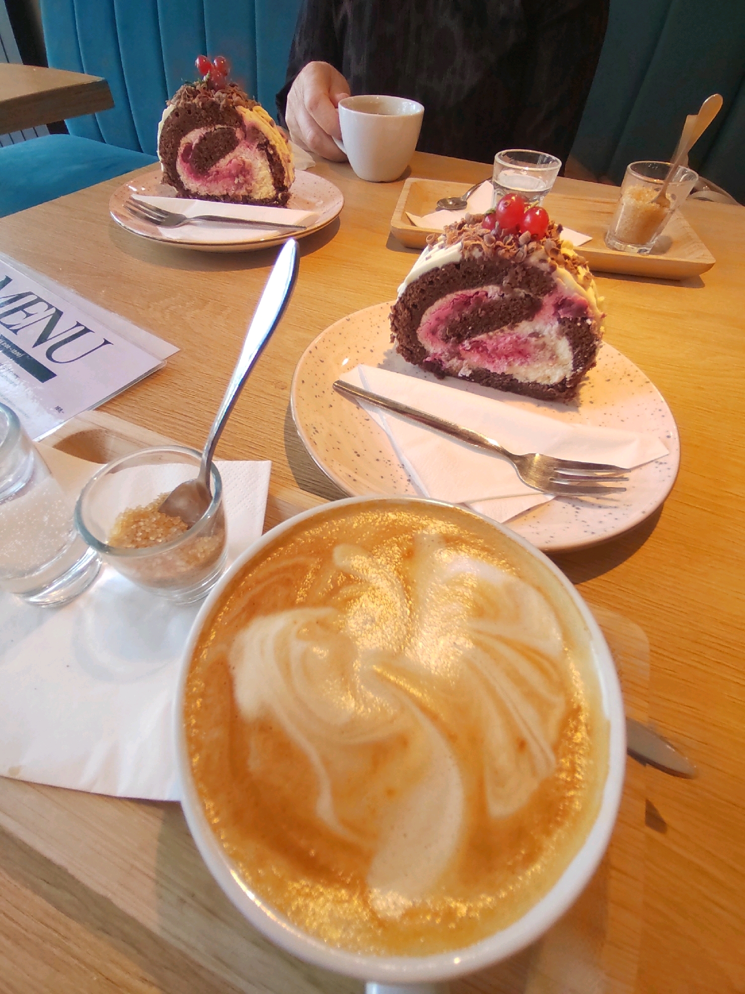 The image depicts a table setting in what appears to be a cafe. In the foreground, a white mug filled with coffee with latte art is positioned to the right. To the left of the coffee, a glass dish containing brown sugar and a small spoon is visible. Behind this, a salt shaker with a silver lid is present.
Further back on the table, a slice of chocolate swiss roll cake topped with white frosting and two red berries rests on a patterned plate, accompanied by a fork. Adjacent to this plate is a folded white napkin. Beyond the cake, a person's hand is seen holding a white cup.
In the background, a bluish upholstered bench seat is noticeable. A wooden table and a menu card on the left complete the scene. The lighting suggests an indoor environment, possibly a cafe. There are no distinct landmarks visible that would pinpoint the exact location in Prague, Czechia.