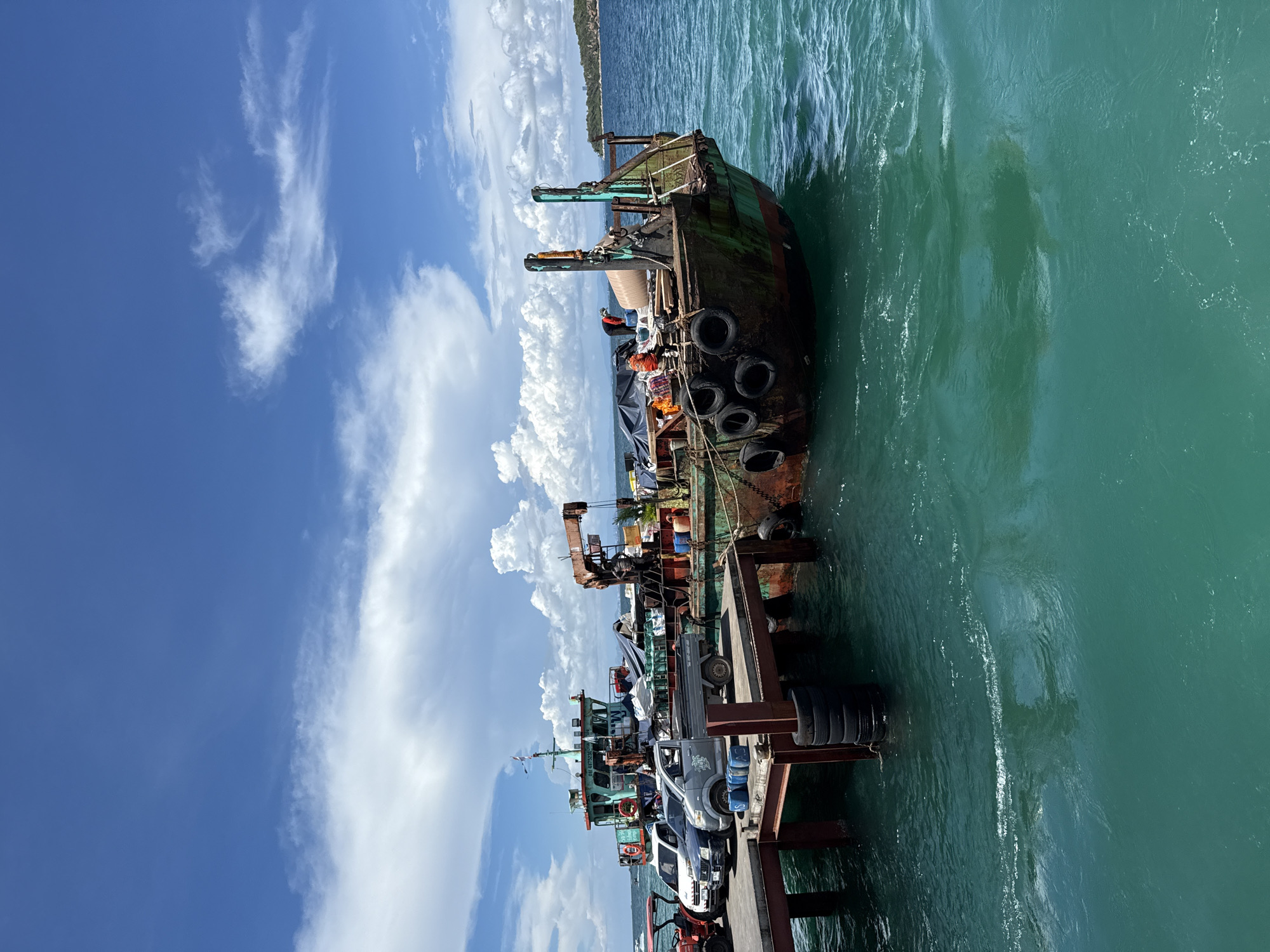 This image captures a scene at a dock in Pattaya City, Thailand. The focus is on several ships and a pier with vehicles parked along it. A dilapidated, rusted ship is prominently docked, featuring numerous tires along its hull. Further along the pier are other smaller ships, accompanied by a selection of cars and a red tractor. The environment includes a clear blue sky with scattered white clouds, contrasted with the turquoise-colored ocean water. The image is taken from a high-angle perspective.