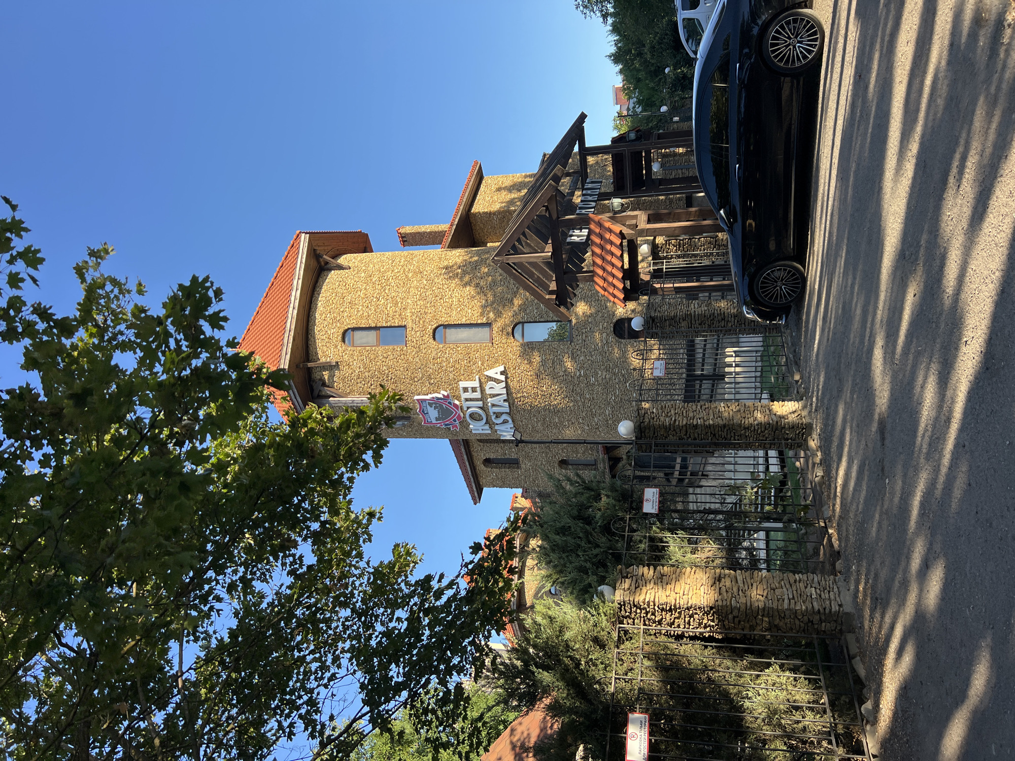 In the city of Fontanka, Ukraine, a black Mercedes-Benz S-Class sedan is parked on a gravel driveway in front of a stone-built hotel with a red tiled roof. The hotel features a prominent sign that reads "HOTEL MASTARA" in large white letters, accompanied by a crest above it. A wrought-iron gate with stone pillars, adorned with two "P" parking signs for restaurant guests, separates the hotel entrance from the driveway. Lush green trees and bushes surround the property, and a large tree with green leaves casts dappled shadows on the ground. The sky is clear and blue.
