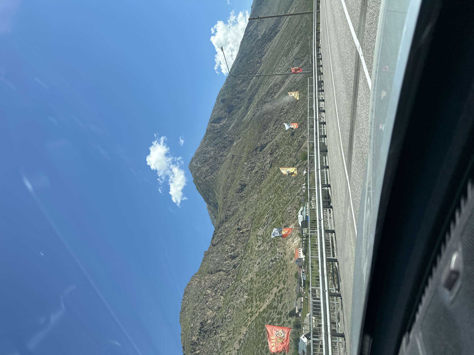 This is a slightly low-angle, eye-level, medium shot taken from inside a vehicle, looking out at a mountainous landscape under a bright blue sky with scattered white clouds. The foreground shows the car's dashboard and windshield, with a sliver of reflection visible on the dashboard. The middle ground features a paved road with white lane markings, bordered by a metal guardrail. Beyond the guardrail, there is a small village with houses and some vegetation. Several flags are planted along the roadside, some featuring red and yellow colors and possibly emblems or text. The background is dominated by a large, green, and rocky mountain that rises steeply into the sky. The overall scene depicts a scenic view of a rural mountainous area in Upper Baksan, Russia.