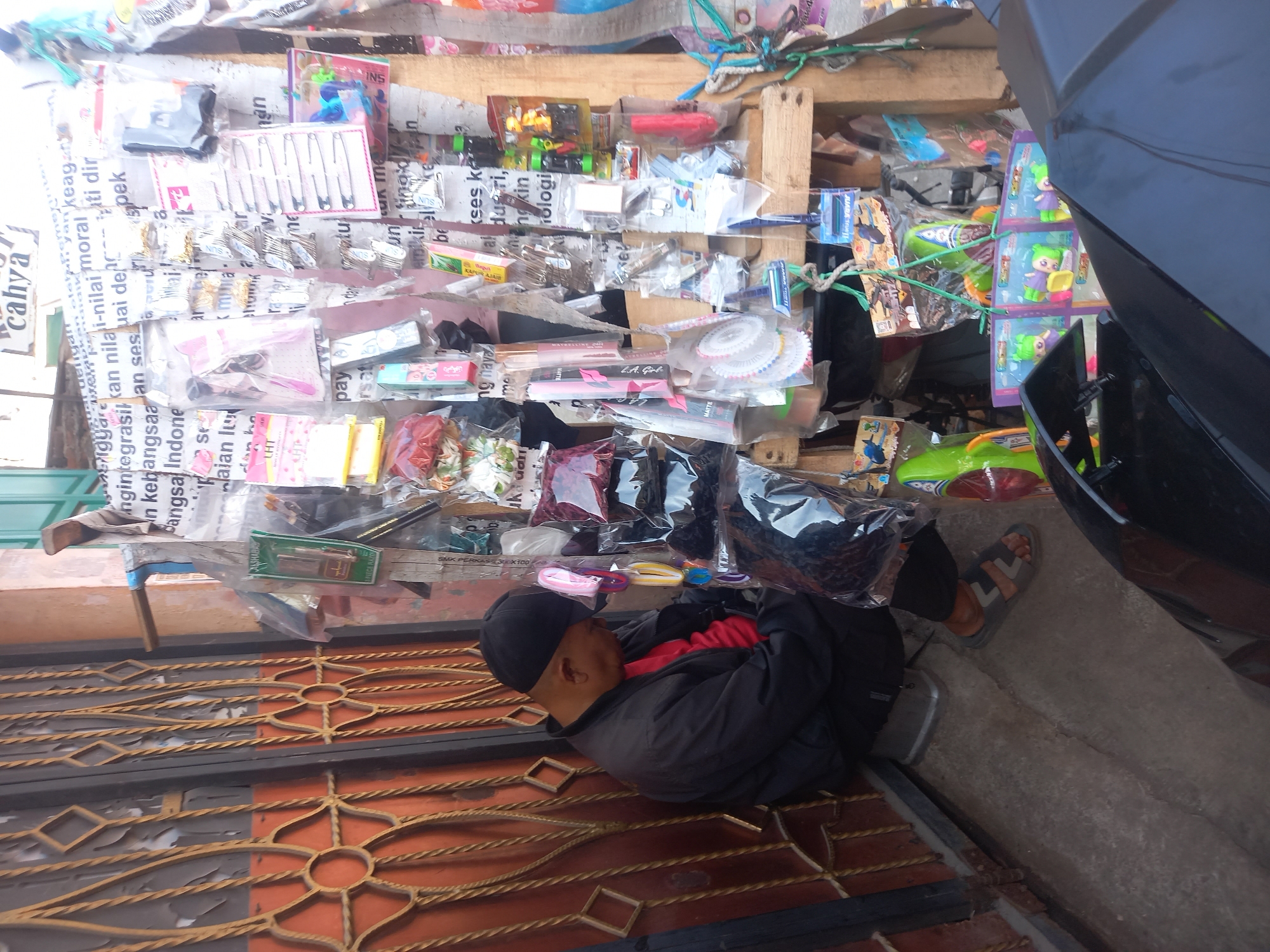 A man wearing a black jacket, red shirt, and black baseball cap is sitting on a low stool next to a storefront. He is facing right and appears to be asleep or resting, with his head down. In front of him, to his right, is a street stall displaying various small items for sale, including hair accessories, toys, and packaged goods. The stall is constructed from wood and paper, with items hanging from wires and hooks. Behind the man and the stall is a metal gate with an ornate design. The ground is paved with concrete. The scene appears to be in a street market or commercial area. The location is identified as Linggar, Indonesia.