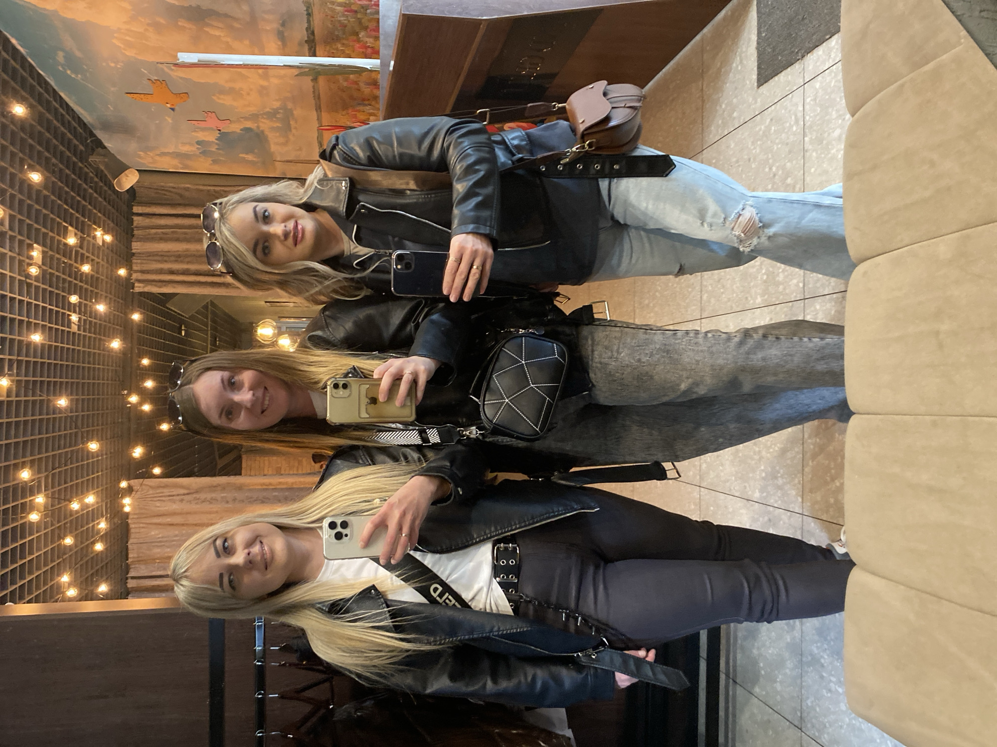 Three women are posing for a picture in what appears to be a mirror. The location seems to be the restaurant "Nebesniy Edinorog" in Chaikovsky, Russia, as indicated by background elements. The women are dressed in similar styles, wearing leather jackets. One woman is wearing ripped jeans, while the other two are wearing darker-colored pants. They are all holding smartphones, seemingly taking a selfie. Behind them is a wall with decorations.
