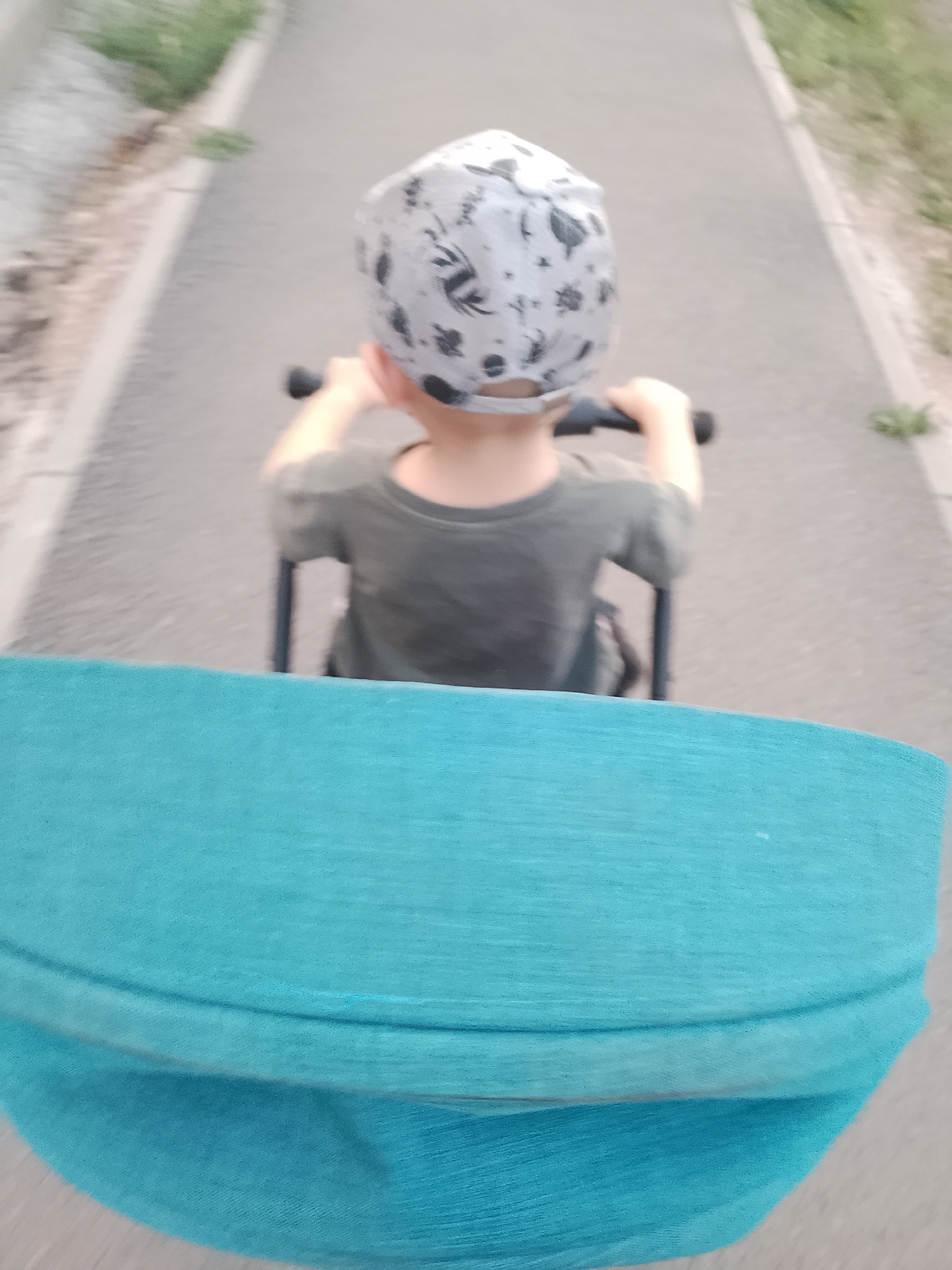 An out-of-focus, medium-angle shot shows a young child wearing a grey and white patterned cap and a grey t-shirt, seen from behind. The child is seated in a stroller and holding onto the stroller's handlebars. The stroller's bright teal fabric is prominently featured in the foreground, obscuring the lower portion of the image. The background depicts a paved pathway with green grass and vegetation on either side, suggesting an outdoor setting in a city or park. The overall blurriness indicates movement, possibly the stroller being pushed.