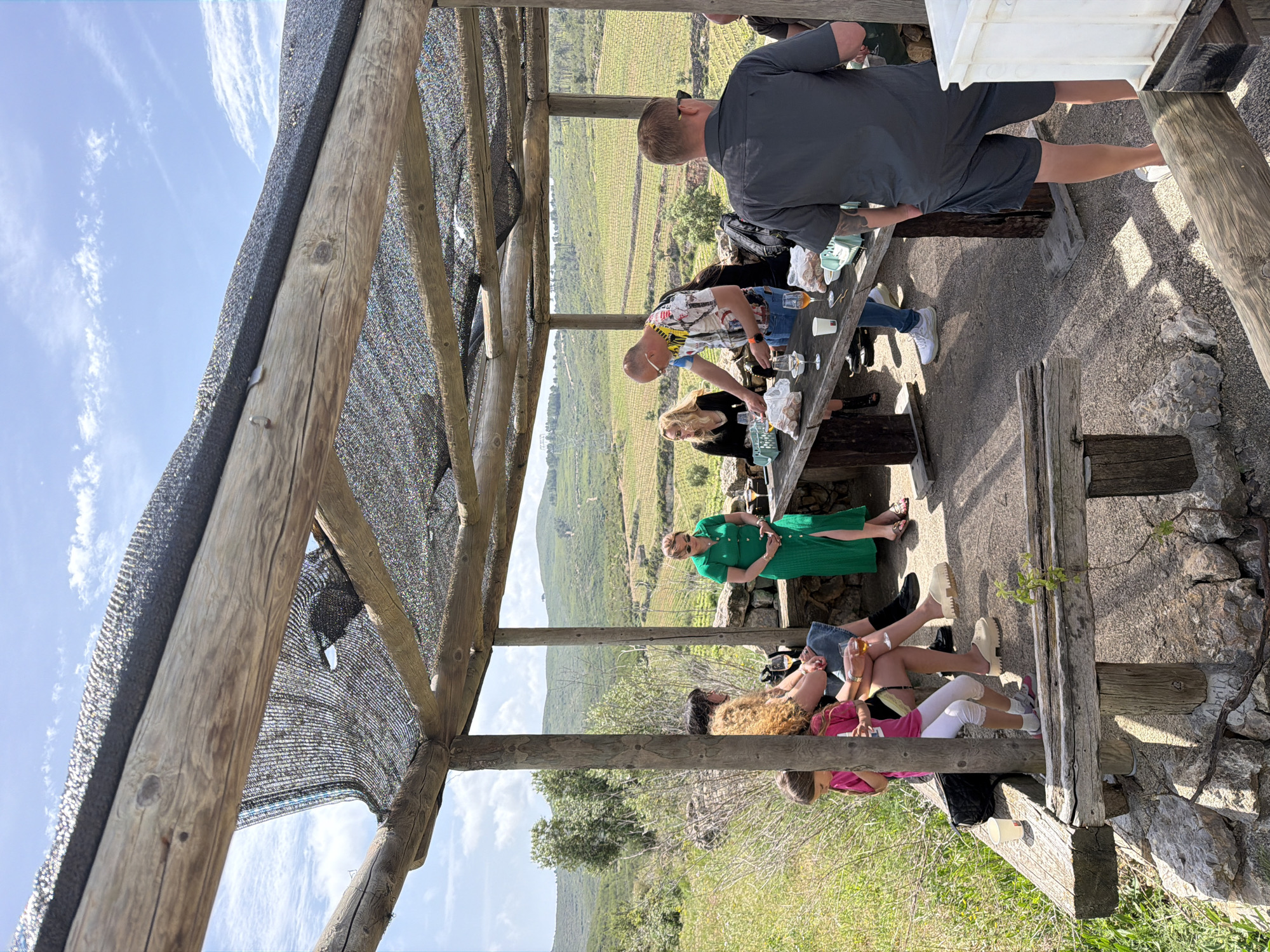People are gathered under a wooden pergola structure, possibly in Bonastre, Spain. They sit around a wooden table with drinks and food. A man in a gray shirt stands at the table, while others are seated. Beyond the structure is a view of rolling hills covered in vineyards. The weather appears sunny. The setting suggests a vineyard or outdoor tasting area.