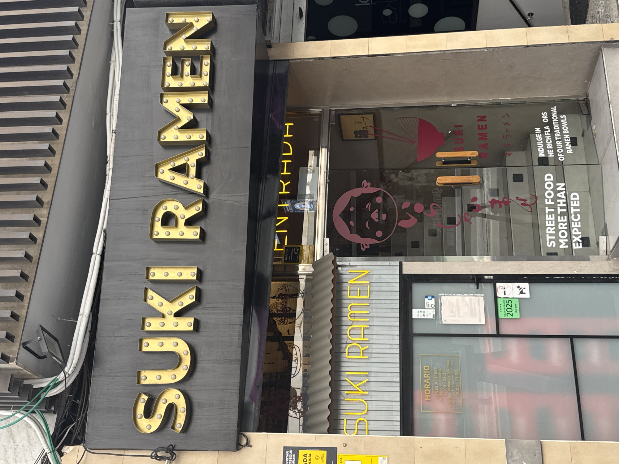 A storefront sign reading "SUKI RAMEN" in large, illuminated golden letters is visible at the top of the image. Below this, a smaller neon sign reads "SUKI RAMEN" in yellow. The entrance to the establishment features glass doors with a frosted lower half. On the left door, a sign displays "HORARIO" (hours) in Spanish, listing opening times for weekdays and weekends. A sticker for "2025 MEJOR VALORADO" (2025 Best Rated) by Uber Eats is affixed to the door. The right glass door has Japanese text and a graphic of a ramen bowl. The interior of the restaurant shows stairs leading up. Text on the glass reads "STREET FOOD MORE THAN EXPECTED" and "INDULGE IN THE RICH FLAVORS OF OUR TRADITIONAL RAMEN BOWLS." The building appears to be in an urban setting, possibly a commercial street. The overall impression is of a modern ramen restaurant.