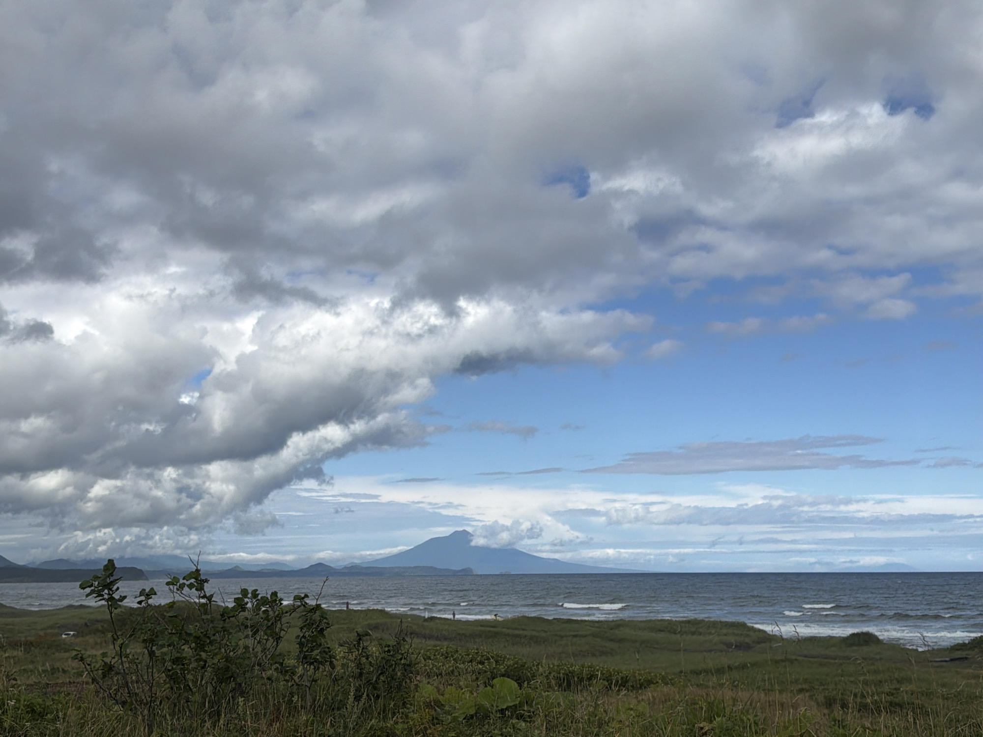 A wide shot captures a scenic coastal landscape in Yuzhno-Kurilsk, Russia. The foreground is dominated by grassy dunes and dense green shrubbery, with tall dry grasses in the foreground. A calm, dark blue ocean stretches across the midground, meeting a sandy beach. In the distance, a large, conical mountain with a visible plume of smoke or clouds erupting from its summit stands against a dramatic sky filled with heavy, grey clouds interspersed with patches of bright blue. The horizon is lined with several mountain ranges, appearing as silhouettes against the sky. The overall atmosphere suggests a potentially overcast or stormy day with a volcanic element present in the landscape.