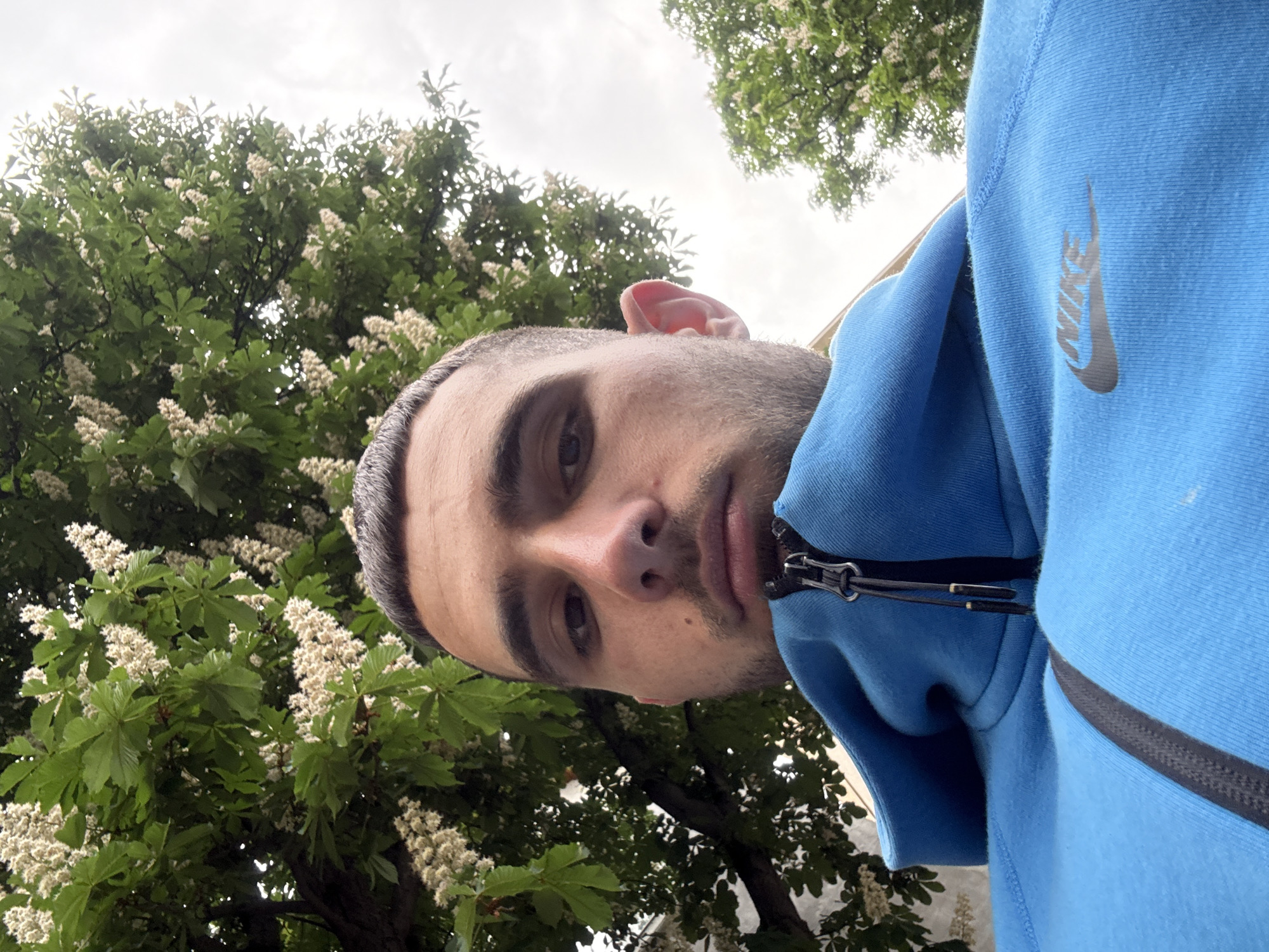 A low-angle, close-up shot features a man with short, dark hair and stubble. He is wearing a blue Nike hoodie with the logo visible. The background is filled with trees with white flowers, suggesting a park or green space. The picture was taken in Sofia, Bulgaria.