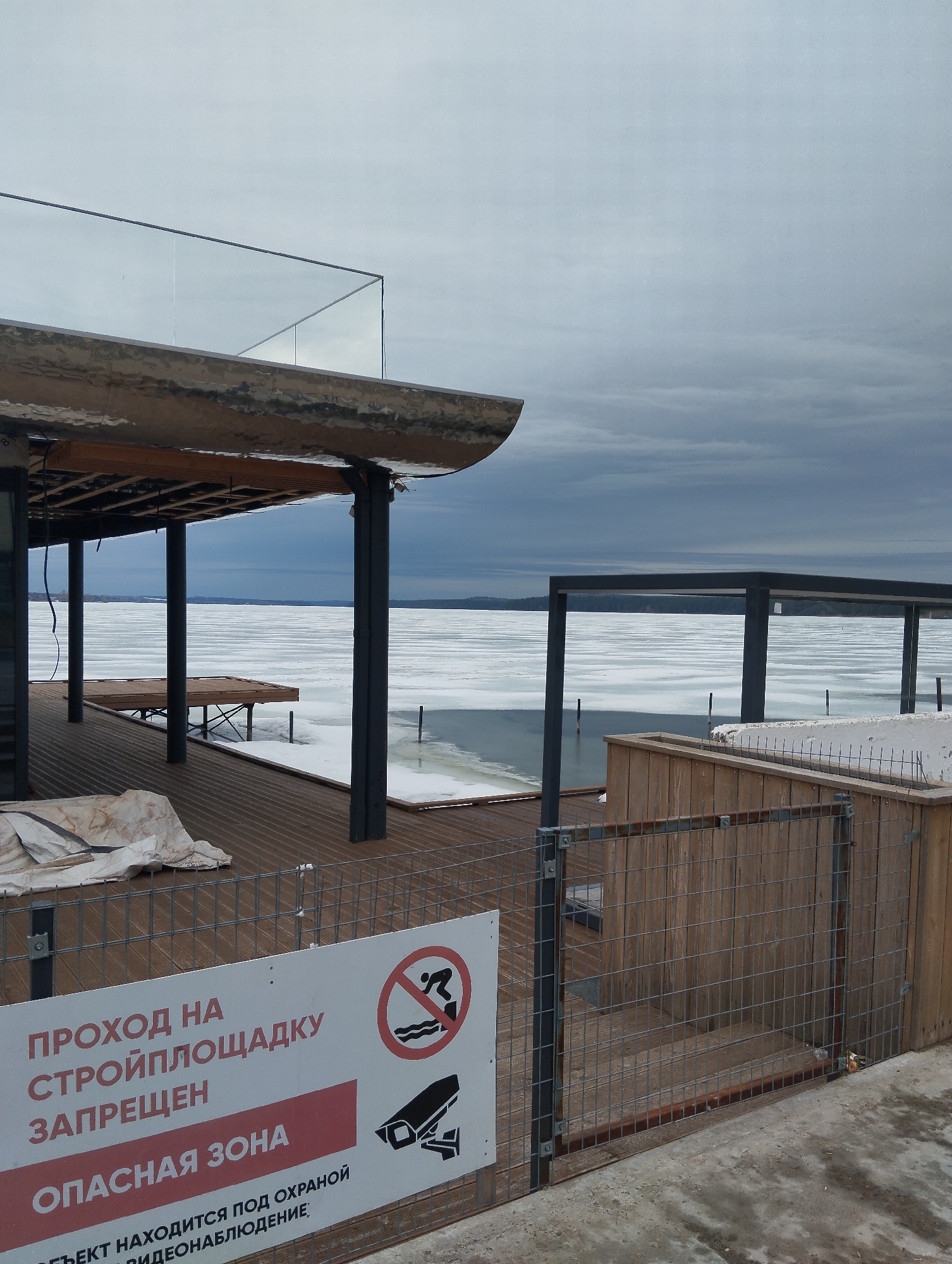 The image depicts a waterfront construction site in Votkinsk, Russia, situated along the Votkinsk Reservoir. The ground is covered with snow and ice, extending to the horizon. On the left is a wooden deck area with a covered structure supported by black pillars. A table stands on the deck, partially covered. A white tarp is visible to the left on the ground. In the foreground, a metal fence with a sign in Russian is evident. The sign indicates that entry to the construction site is prohibited and that the area is under video surveillance. To the right of the fence is a wooden structure, possibly a planter or border. The sky is overcast with grey clouds.