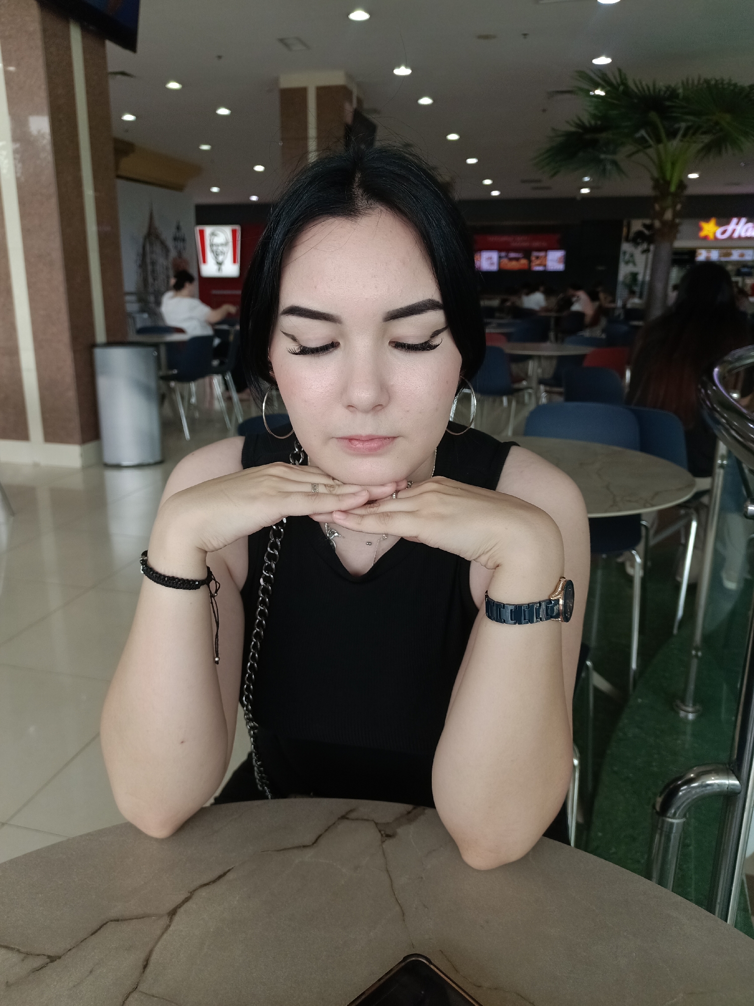 A young woman with dark hair and makeup, wearing a black top and jewelry, sits at a table in an indoor food court. Her eyes are closed, and her hands are resting on her chin. In the background, there are tables, chairs, and food establishments, including a KFC. The location is identified as a food court in Shymkent, Kazakhstan.