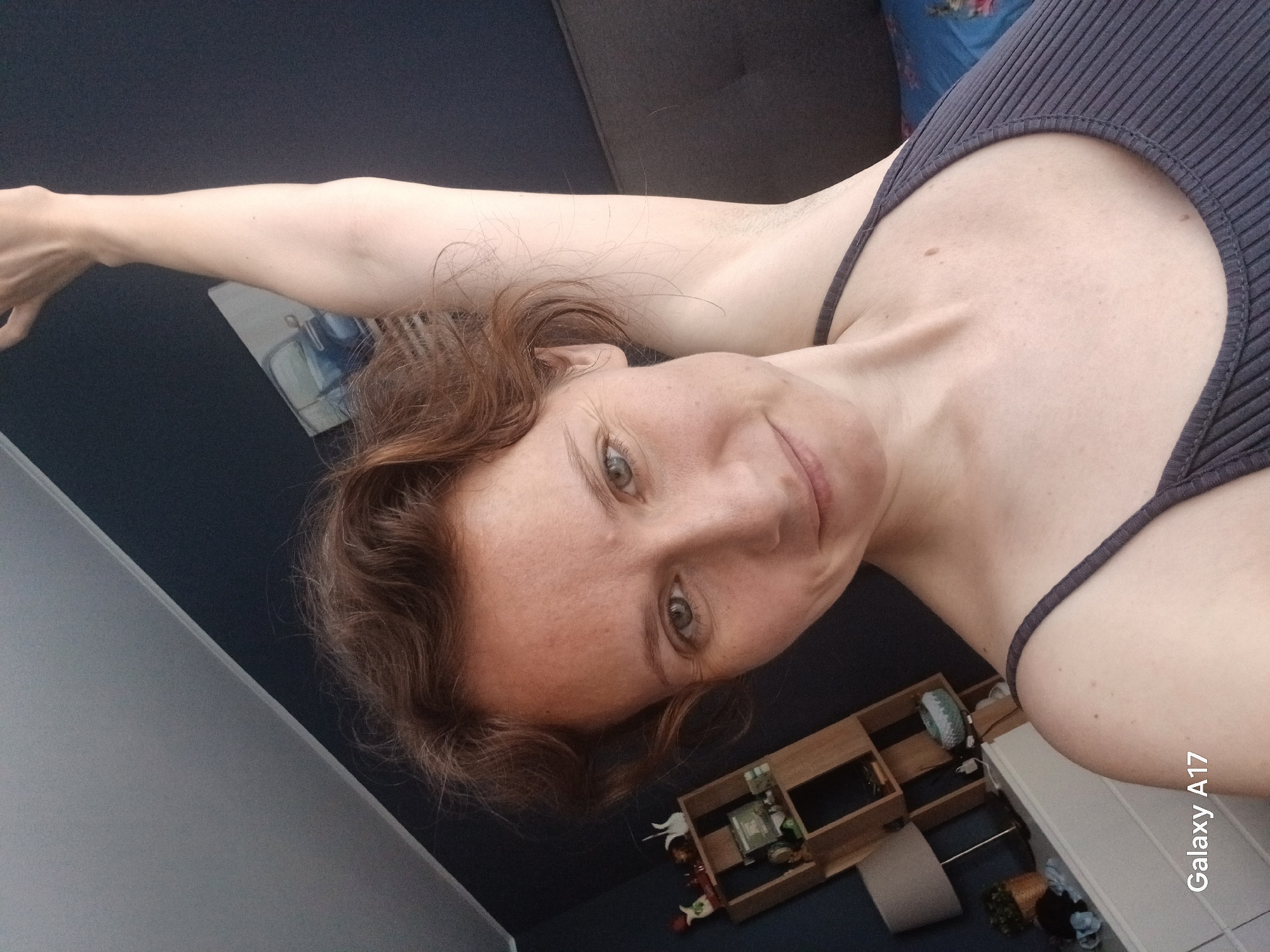 A reddish-haired woman lies down in what appears to be an apartment. She wears a dark-colored, ribbed tank top and is looking into the camera. Her left arm is extended above her head. The room has a dark-colored wall and a light grey wall. A wooden shelf with objects is partially visible near a white baseboard. A portion of a sofa is also visible. The location is unknown and cannot be identified as Yekaterinburg, Russia, from the given image. The lower right corner of the image has the text "Galaxy A17".