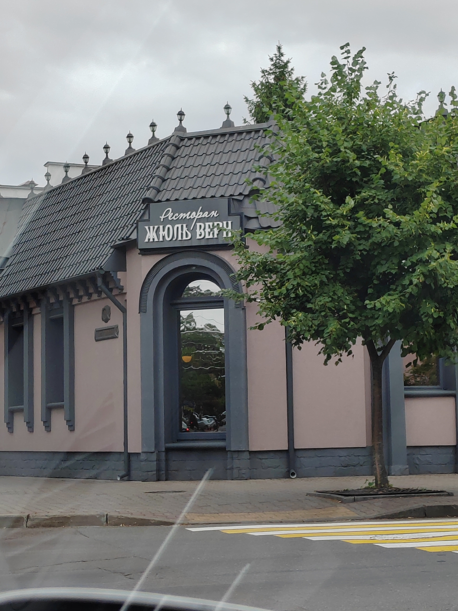 The image shows a street view of a building with a sign that reads "Ресторан ЖЮЛЬ ВЕРН" (Restaurant Jules Verne) in Cyrillic. The building has a gray, tiled roof with decorative lamps and a pink facade with gray architectural details. There is a large arched window reflecting trees and sky. A green tree stands to the right of the building, and a pedestrian crossing is visible in the foreground. The scene appears to be in a city setting, likely Brest, Belarus, given the Cyrillic signage and architectural style. There are no people visible in the image. The sky is overcast.
