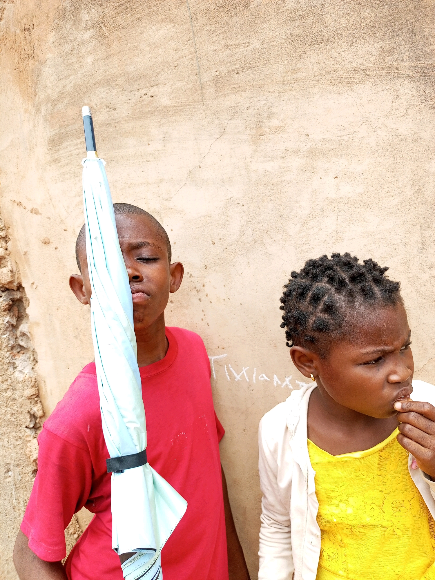 Two children stand near a textured, light brown wall. The child on the left, a boy wearing a red t-shirt, holds a light blue, closed umbrella vertically in front of his face, obscuring his eyes and part of his nose. His eyes are closed and his lips are pursed. The child on the right, a girl wearing a yellow dress and a cream-colored cardigan, has dark, intricately braided hair. She is looking to her right, her right hand touching her mouth. A small gold hoop earring is visible on her right ear. Faint white writing, possibly graffiti, is visible on the wall behind the children. The environment appears to be an outdoor urban setting in Tempure, Nigeria, with the wall suggesting it could be the side of a building.
